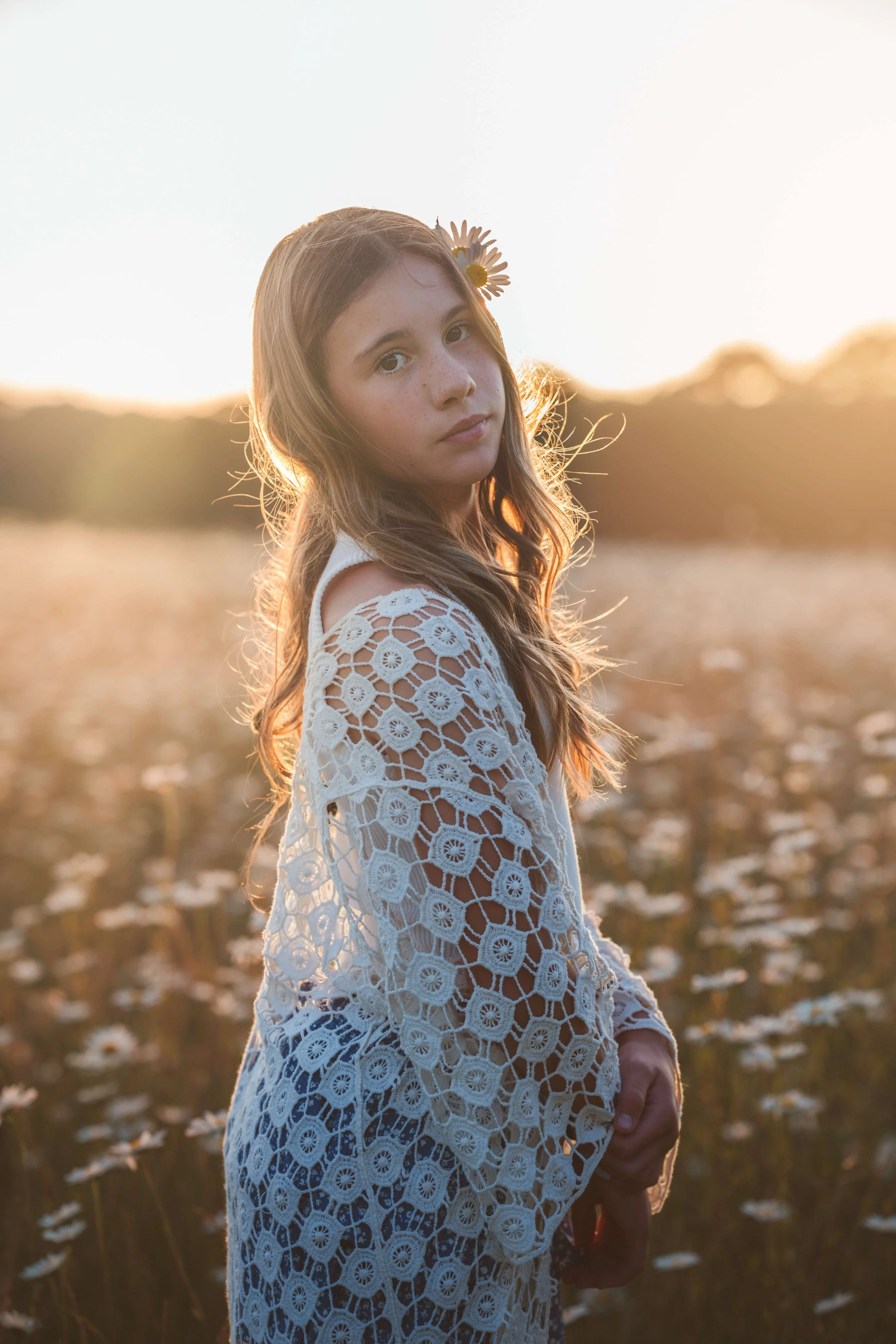 A young girl with long wavy hair and a daisy flower in her hair, standing in a field at sunset, wearing a white lace crochet top.