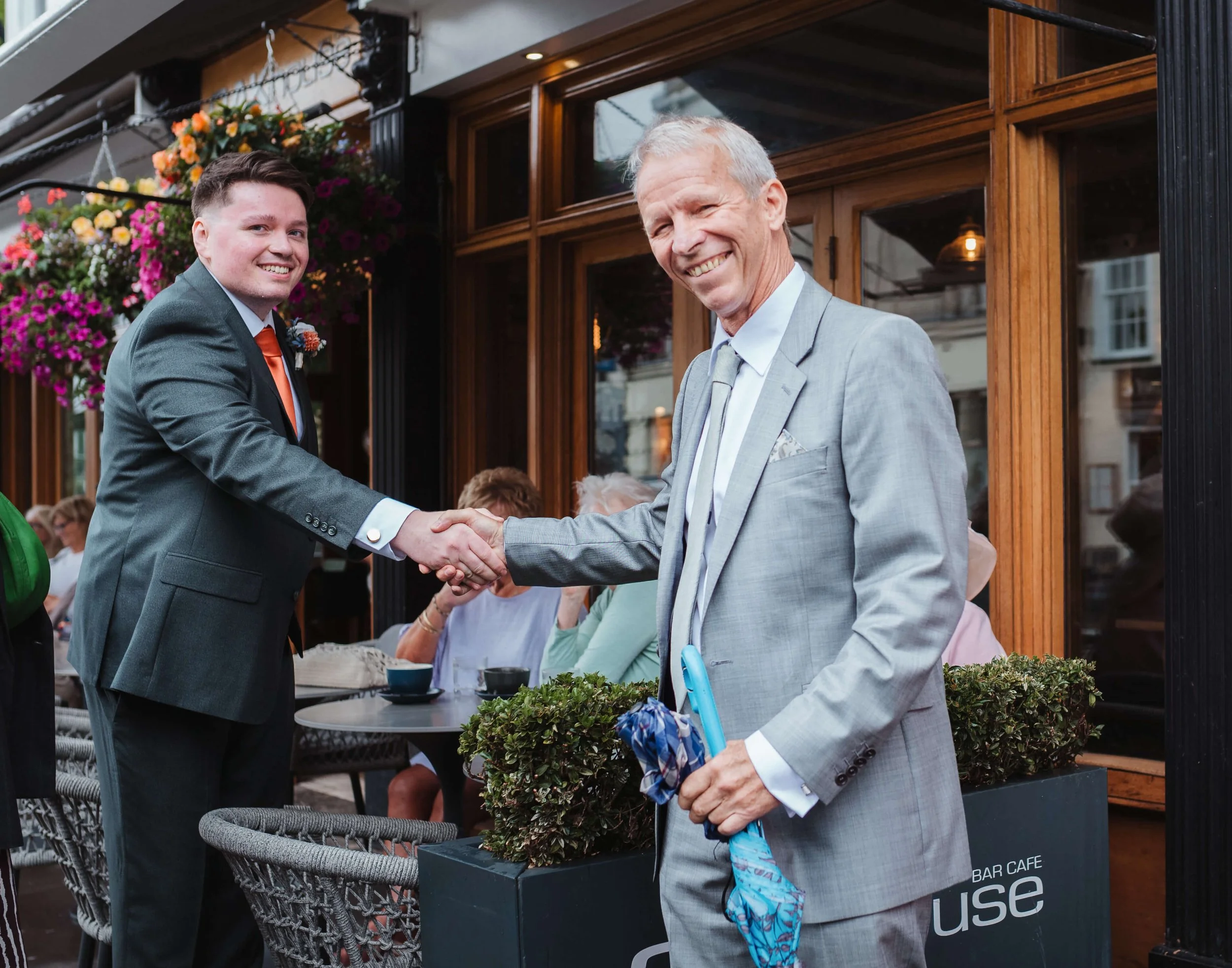 Two men in suits shaking hands outside a restaurant or café with hanging flower baskets. One man is young with dark hair, wearing a dark gray suit and orange tie. The other is older with gray hair, wearing a light gray suit, holding a blue umbrella a