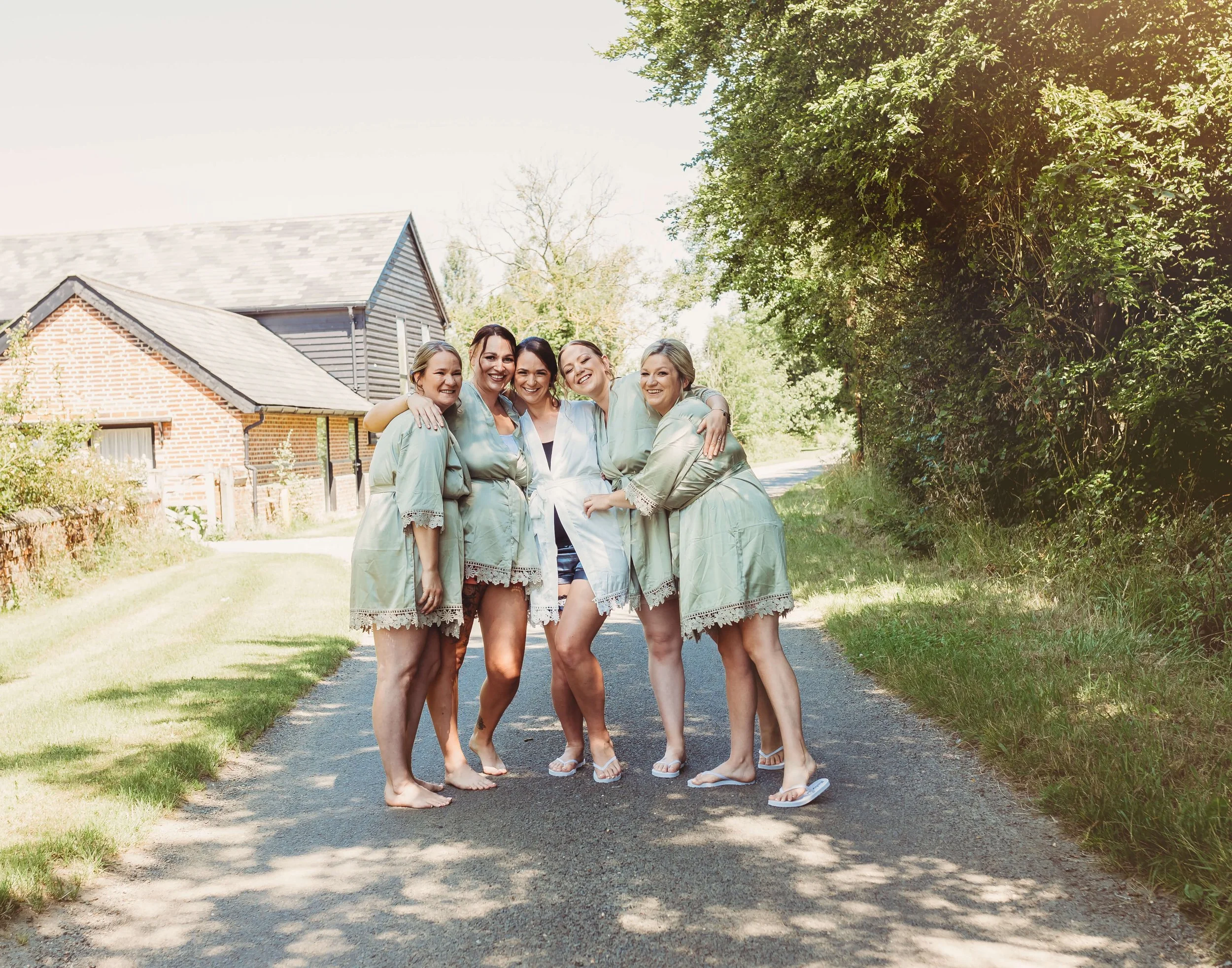 Group of five women standing on a rural road, hugging and smiling, dressed in light green robes and casual clothing, with houses and trees in the background during daytime.