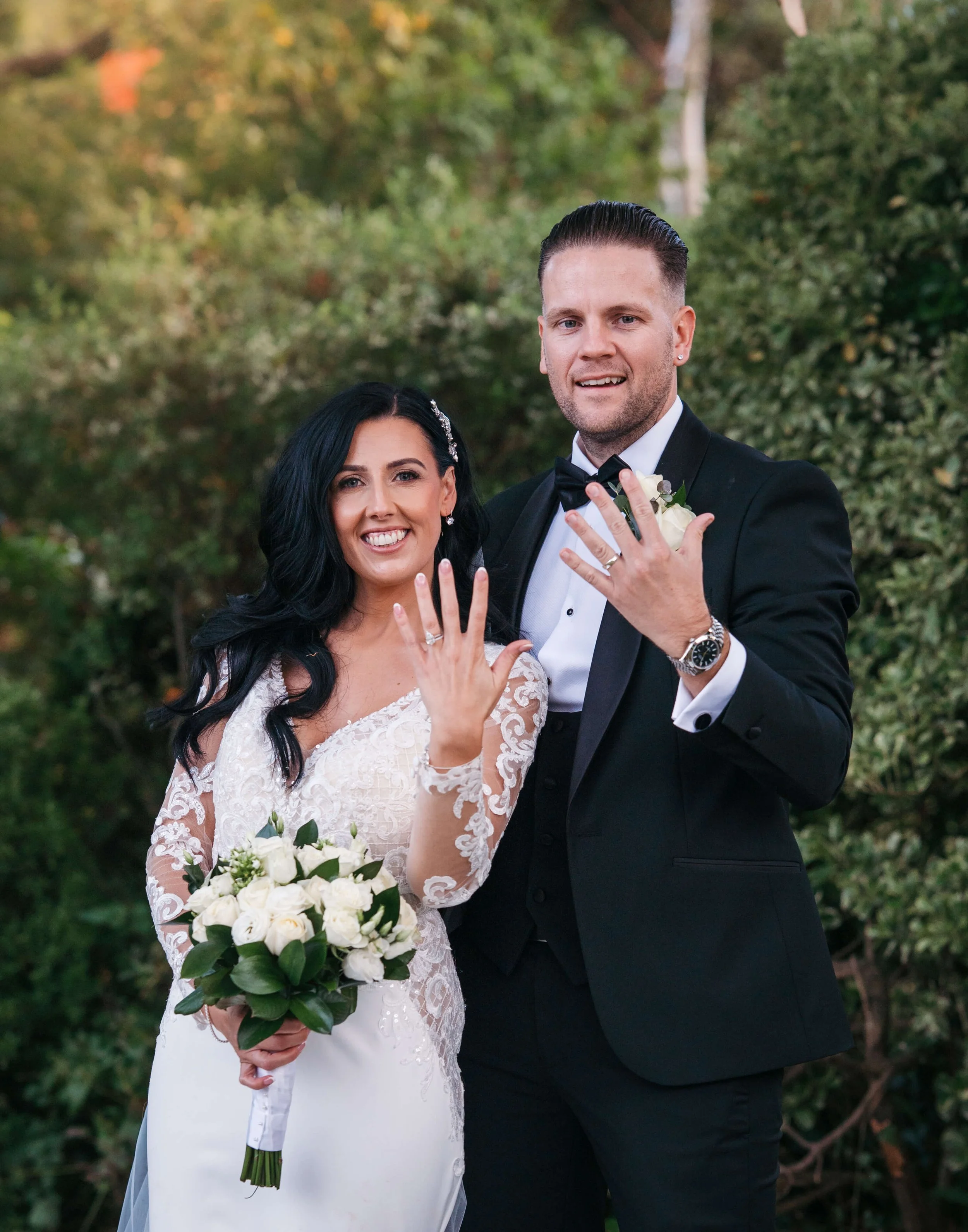 A newlywed couple showing off their wedding rings in an outdoor setting with green foliage in the background.