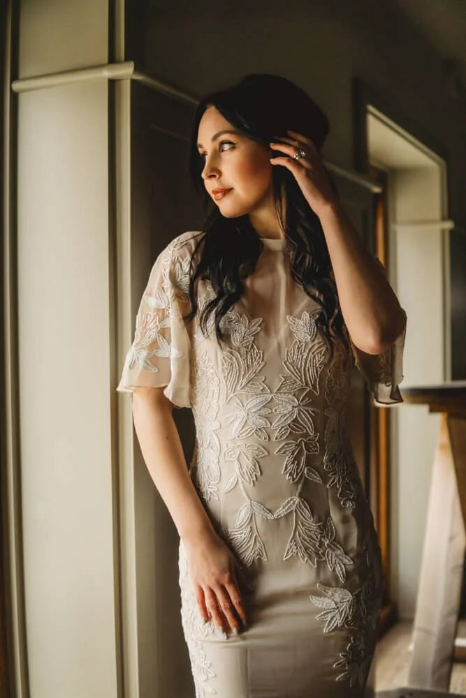 A woman with long black hair wearing a beige dress with floral embroidery, standing near a window and looking to her left.