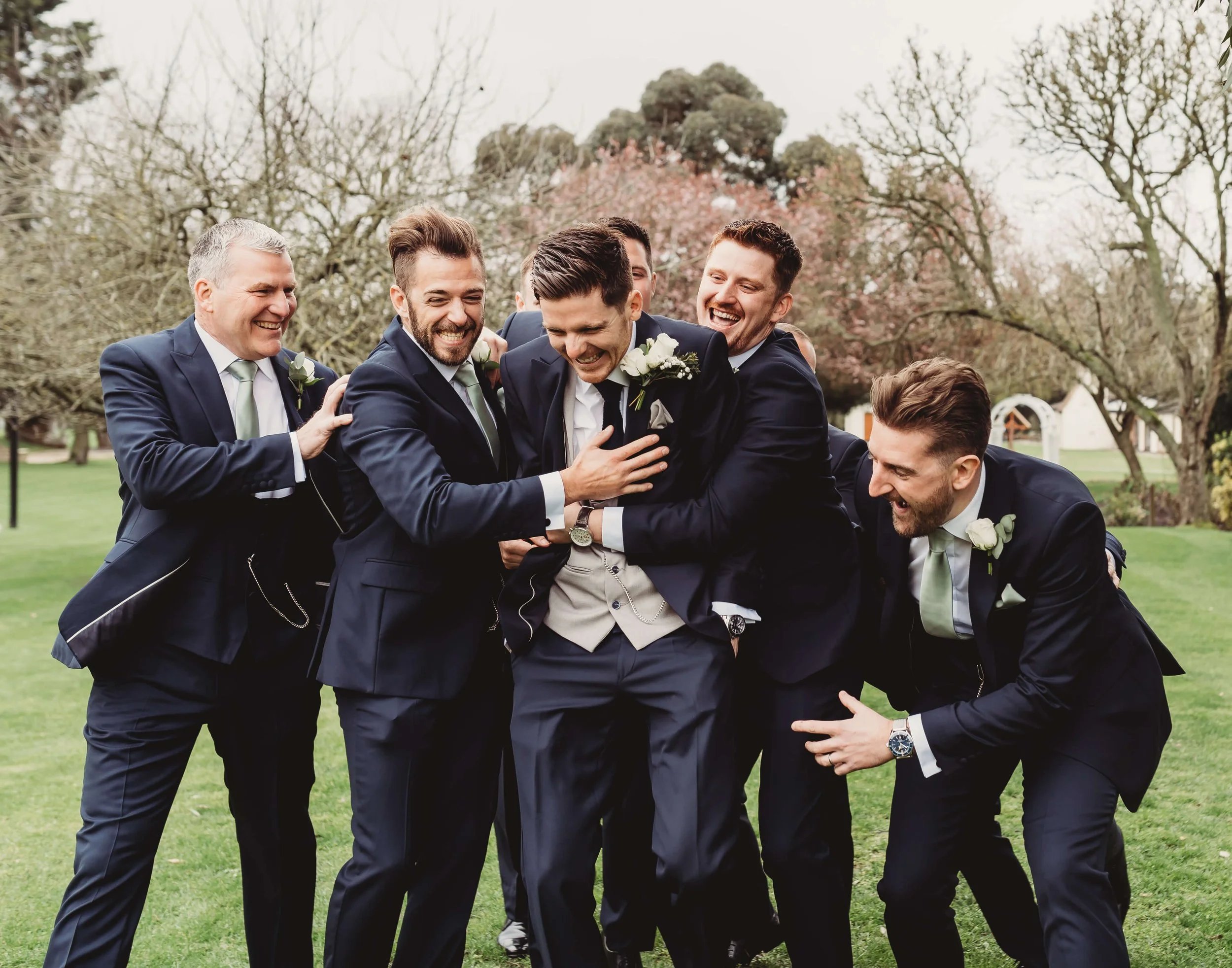 A group of six men in suits having fun and laughing outside during a wedding, with trees and a small building in the background.