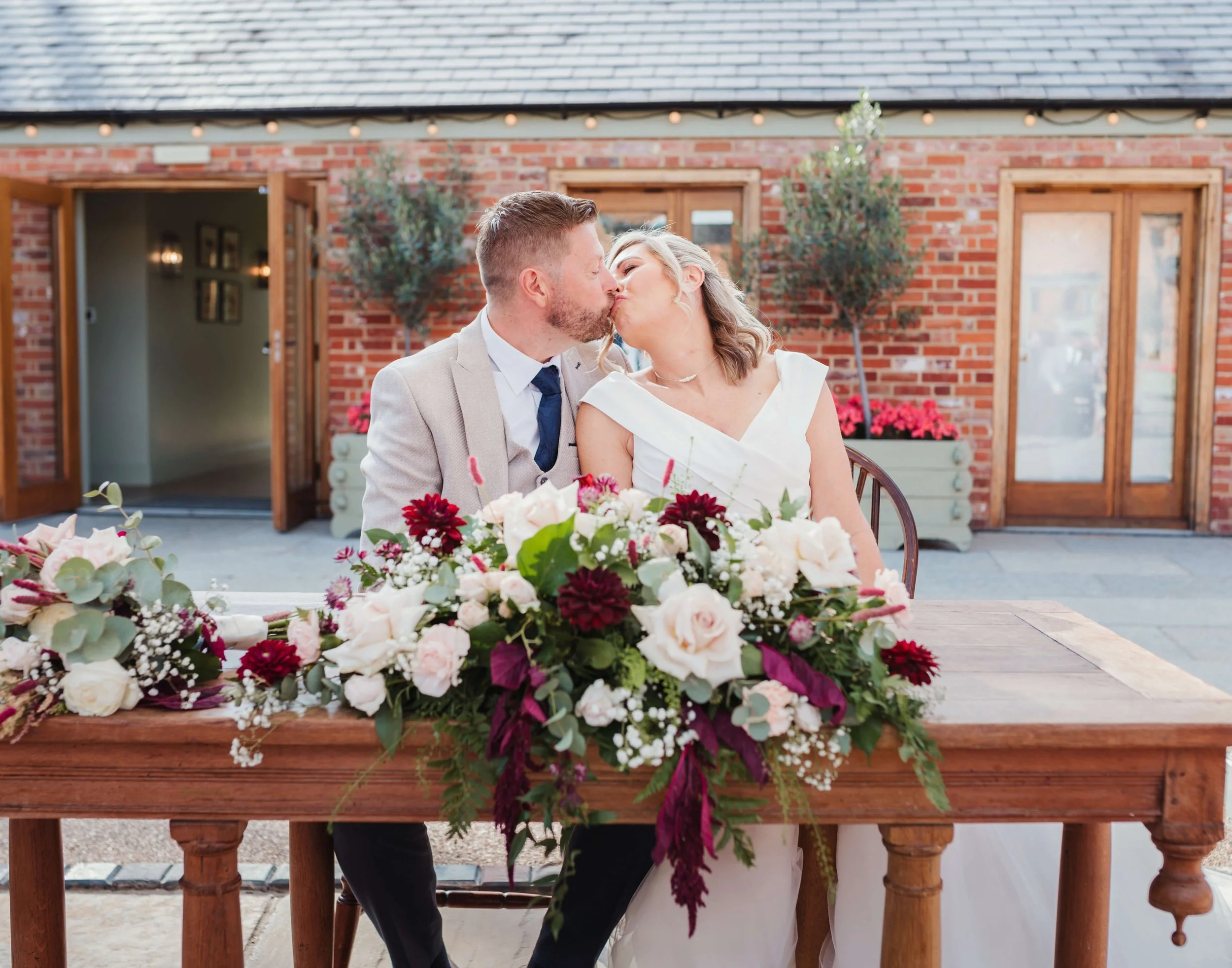 Wedding couple kissing at a decorated table with a large floral centerpiece, outside brick building with open doors and potted plants