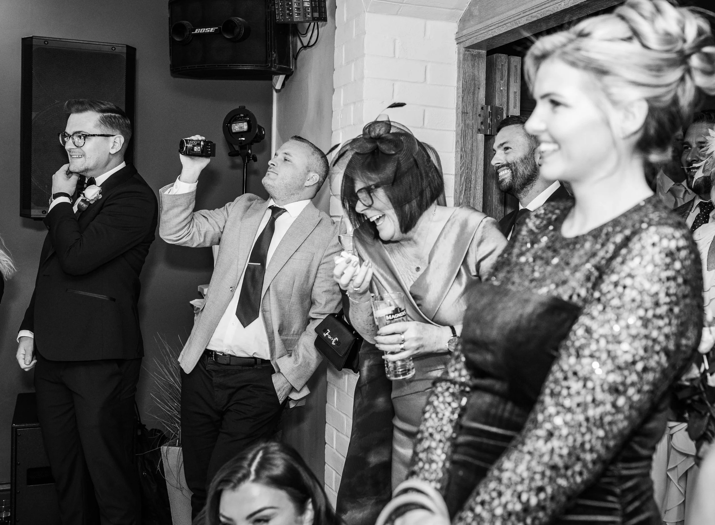 People at a social event, smiling and laughing, including a woman in the foreground with a robbon. Someone is filming the scene with a camcorder.