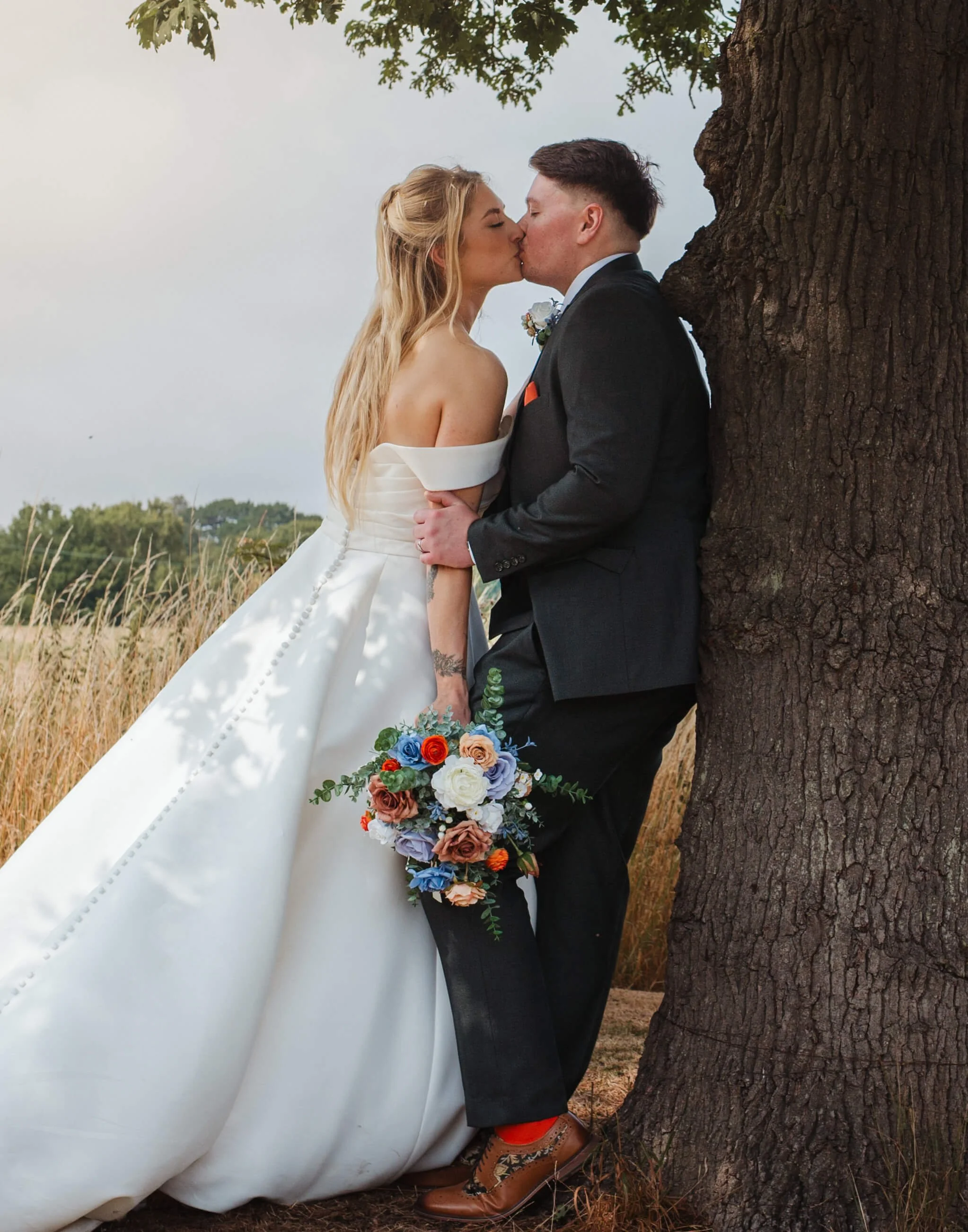 A bride and groom sharing a kiss outdoors, with the bride holding a colorful bouquet, standing next to a large tree in a field.