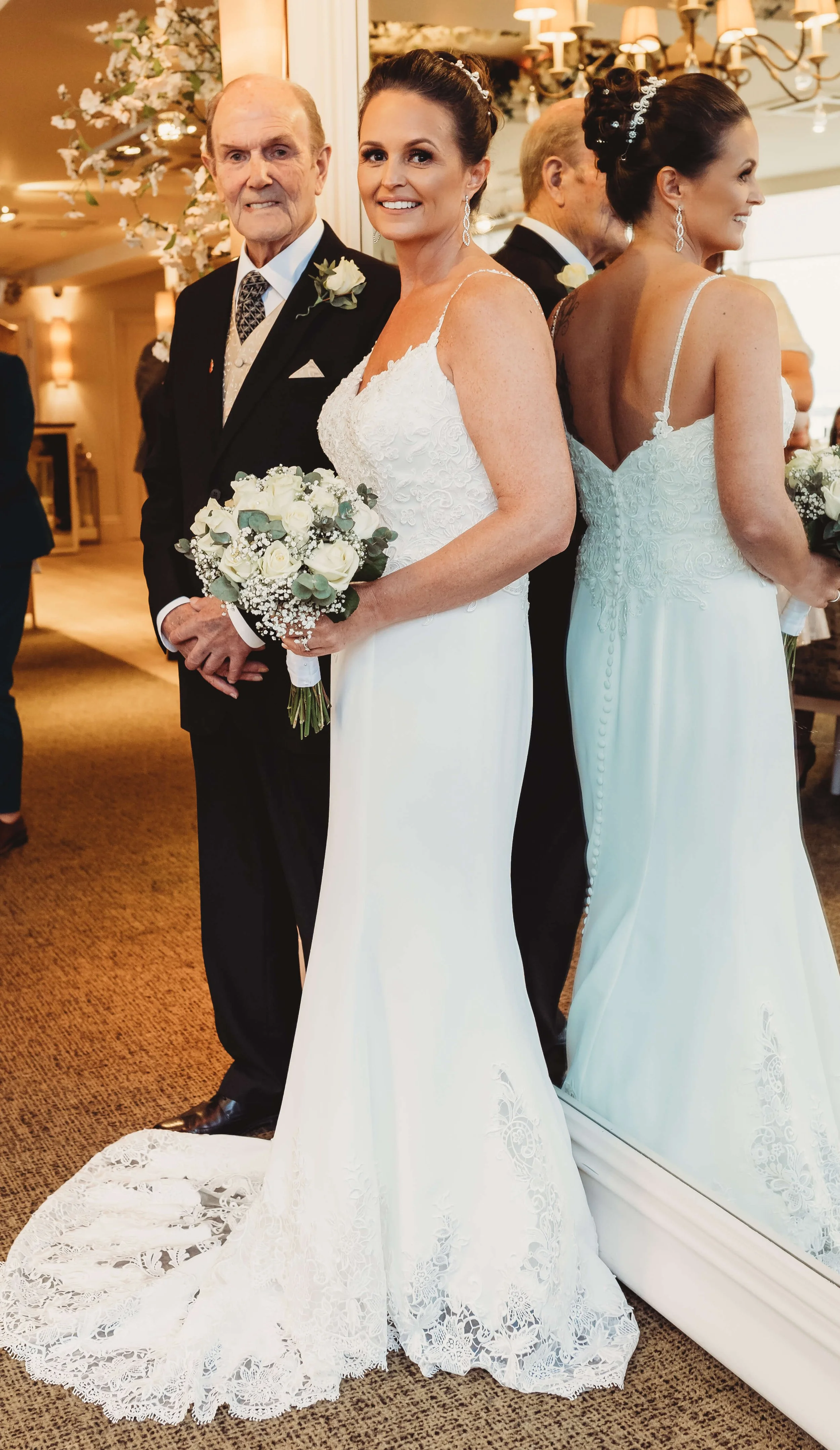A bride in a white wedding dress holding a bouquet standing next to an elderly man in a suit, with a mirror reflecting her image in a wedding venue decorated with flowers and chandeliers.