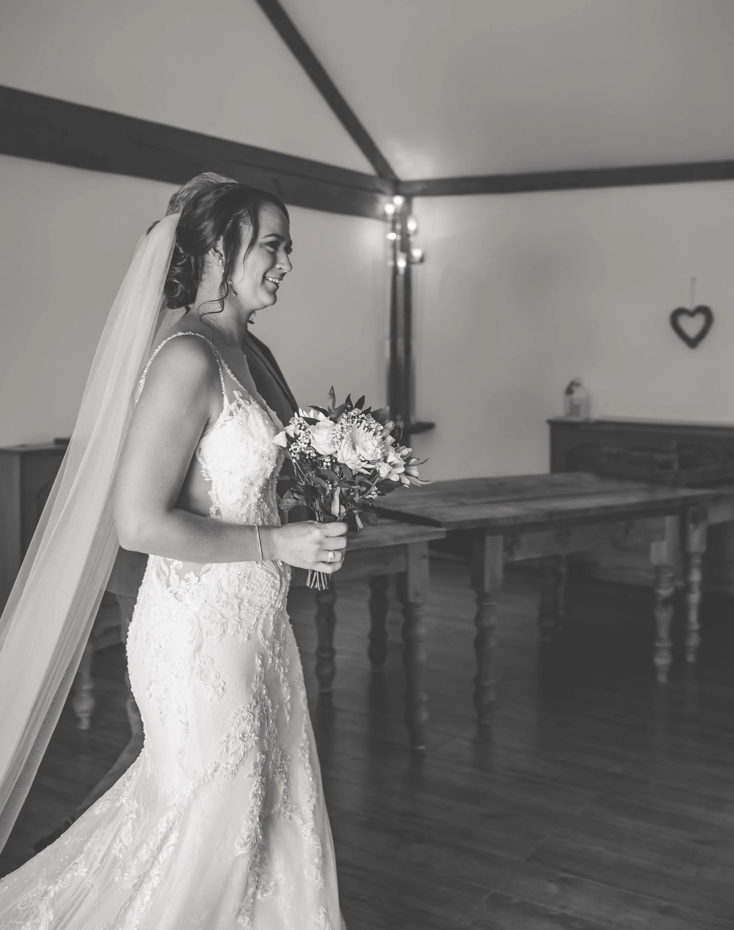 Black and white photo of a smiling bride in a wedding dress holding a bouquet, standing indoors with wooden furniture and a heart-shaped decoration on the wall.
