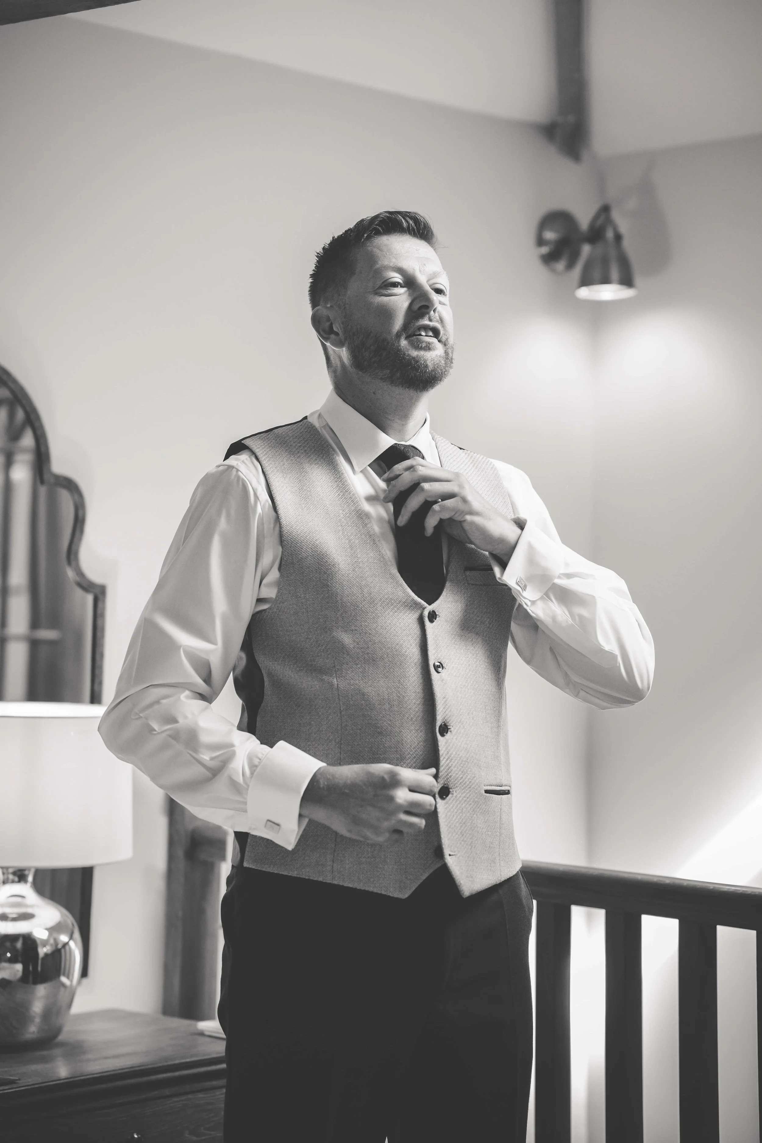 A man in formal attire adjusts his tie, standing indoors, under a wall-mounted light fixture.
