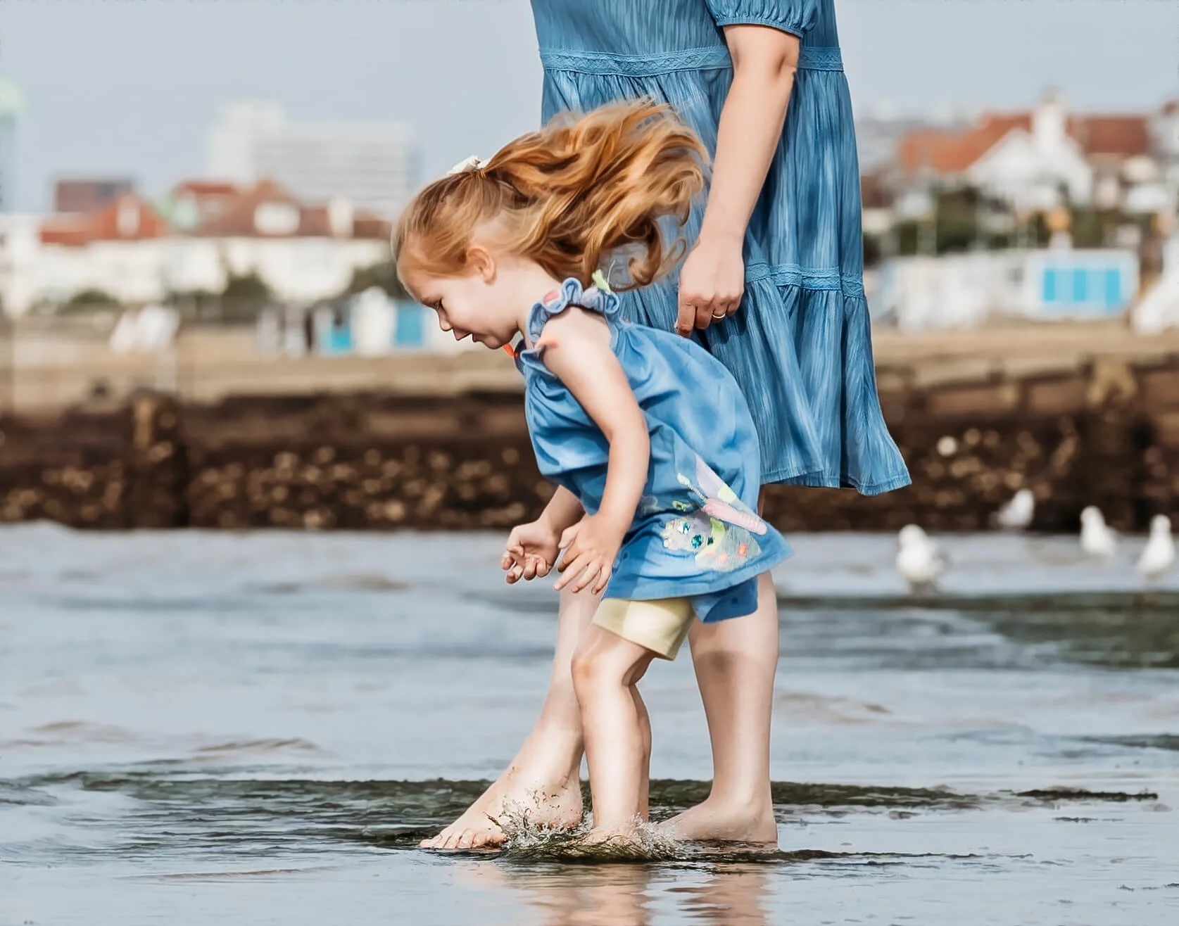 A young girl with red hair and an adult woman, possibly her mother, are barefoot and at the beach with their feet in the water. The girl is bending down, laughing or smiling, with her hair flowing. They are both wearing blue dresses. The background s