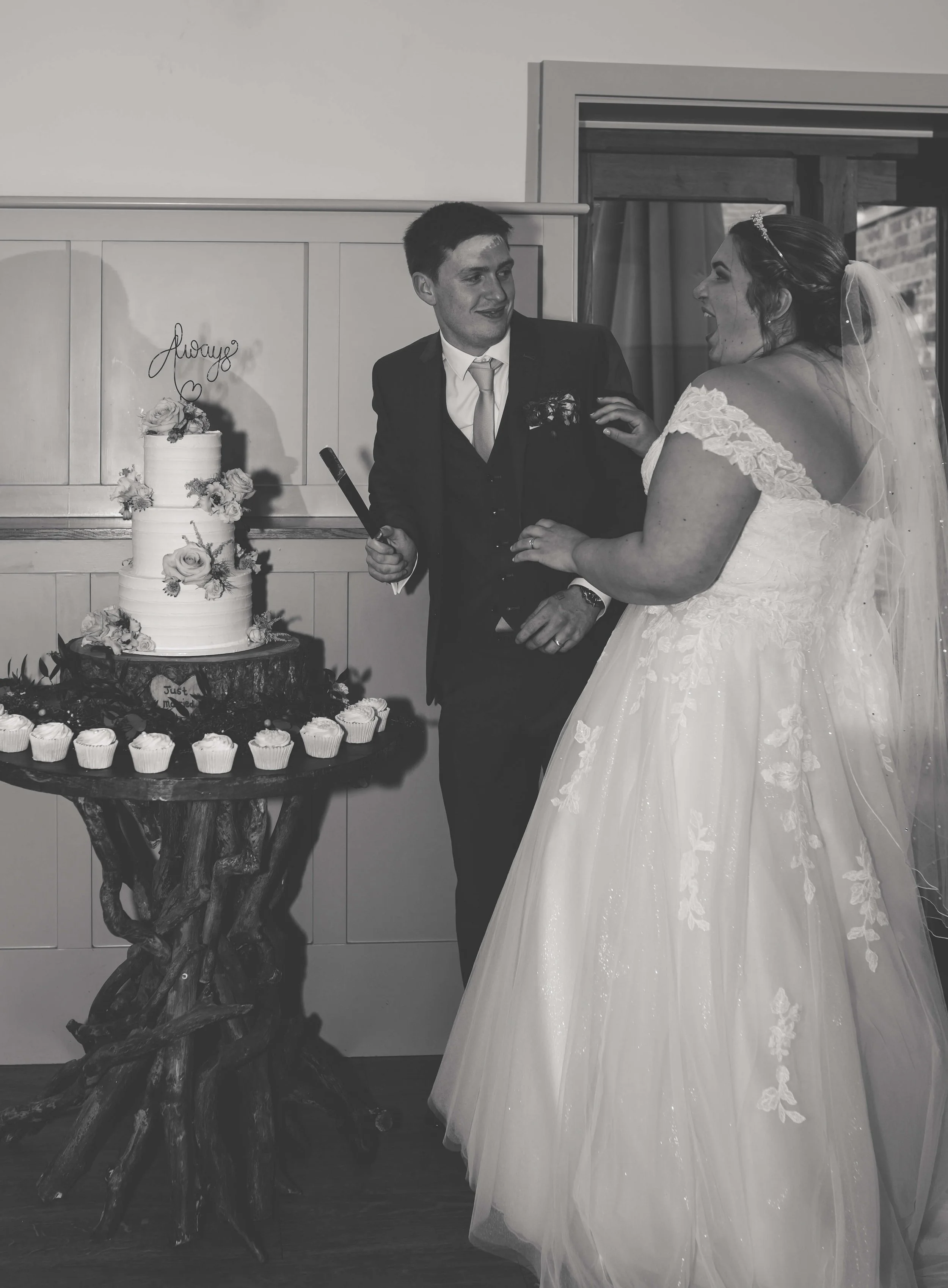 A bride and groom laughing together at their wedding, standing next to a wedding cake with cupcakes on a rustic wooden table.