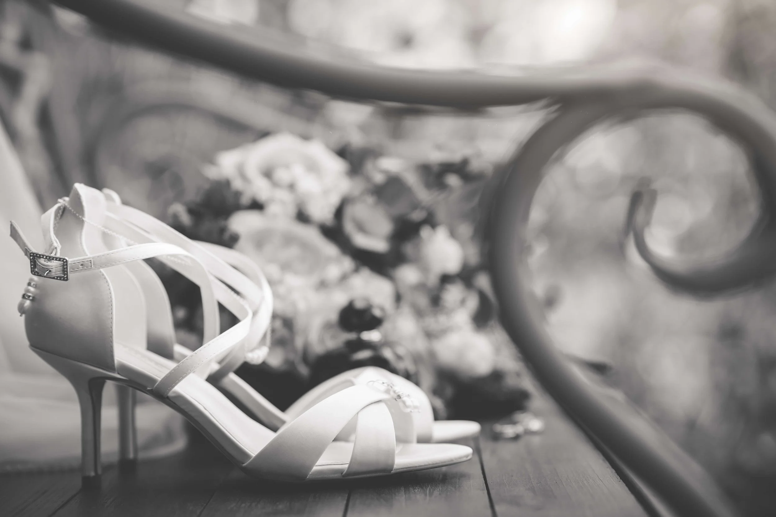 A pair of elegant white high-heeled sandals with straps, placed on wooden flooring. Blurred background showing some decorative elements.