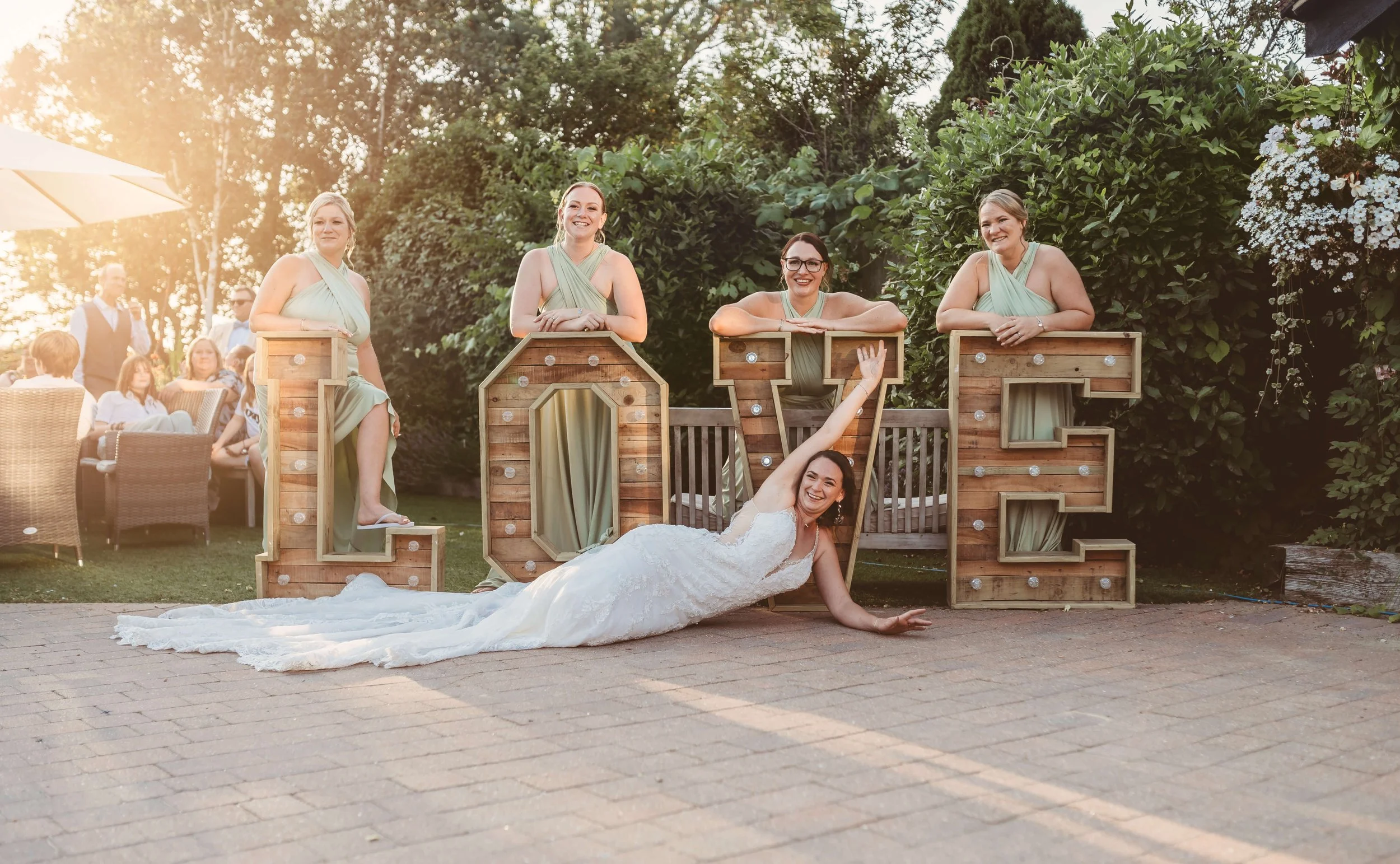Wedding celebration with a bride in a white dress lying on the ground in front of illuminated wooden letters spelling 'LOVE', with four bridesmaids in green dresses sitting and leaning on the letters in an outdoor garden setting.