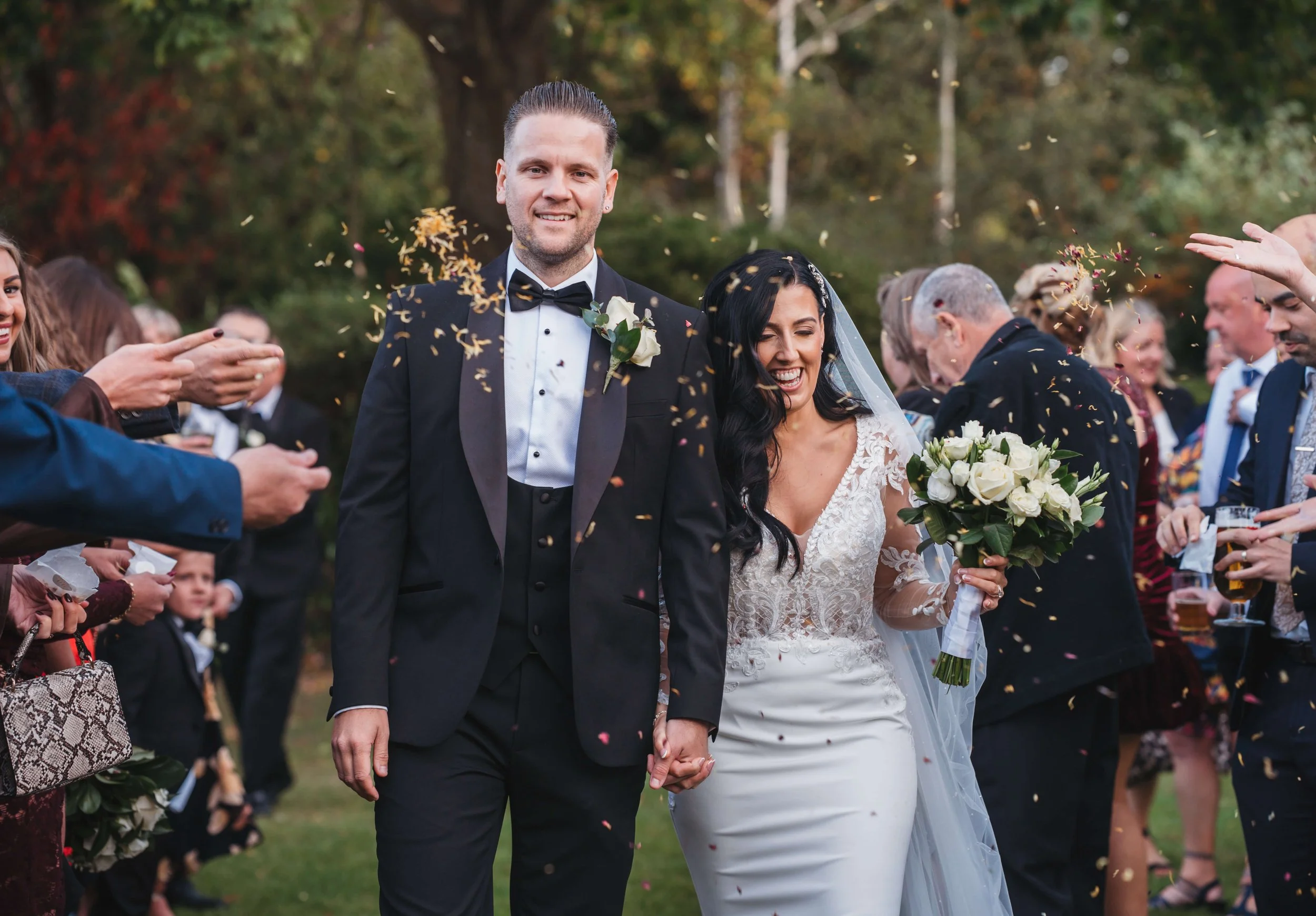 A newlywed couple walking hand in hand while guests celebrate around them with confetti outdoors.