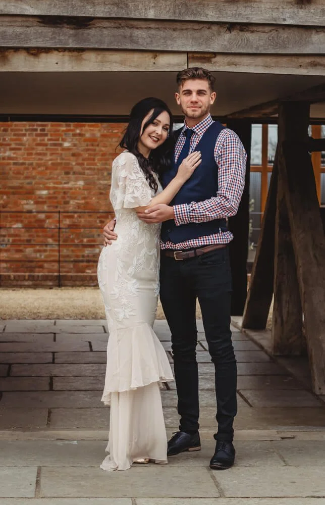 A couple standing close together outdoors, with a brick wall and wooden structure in the background. The woman is wearing a white dress with lace details, and the man is wearing a checkered shirt with a vest and dark pants.