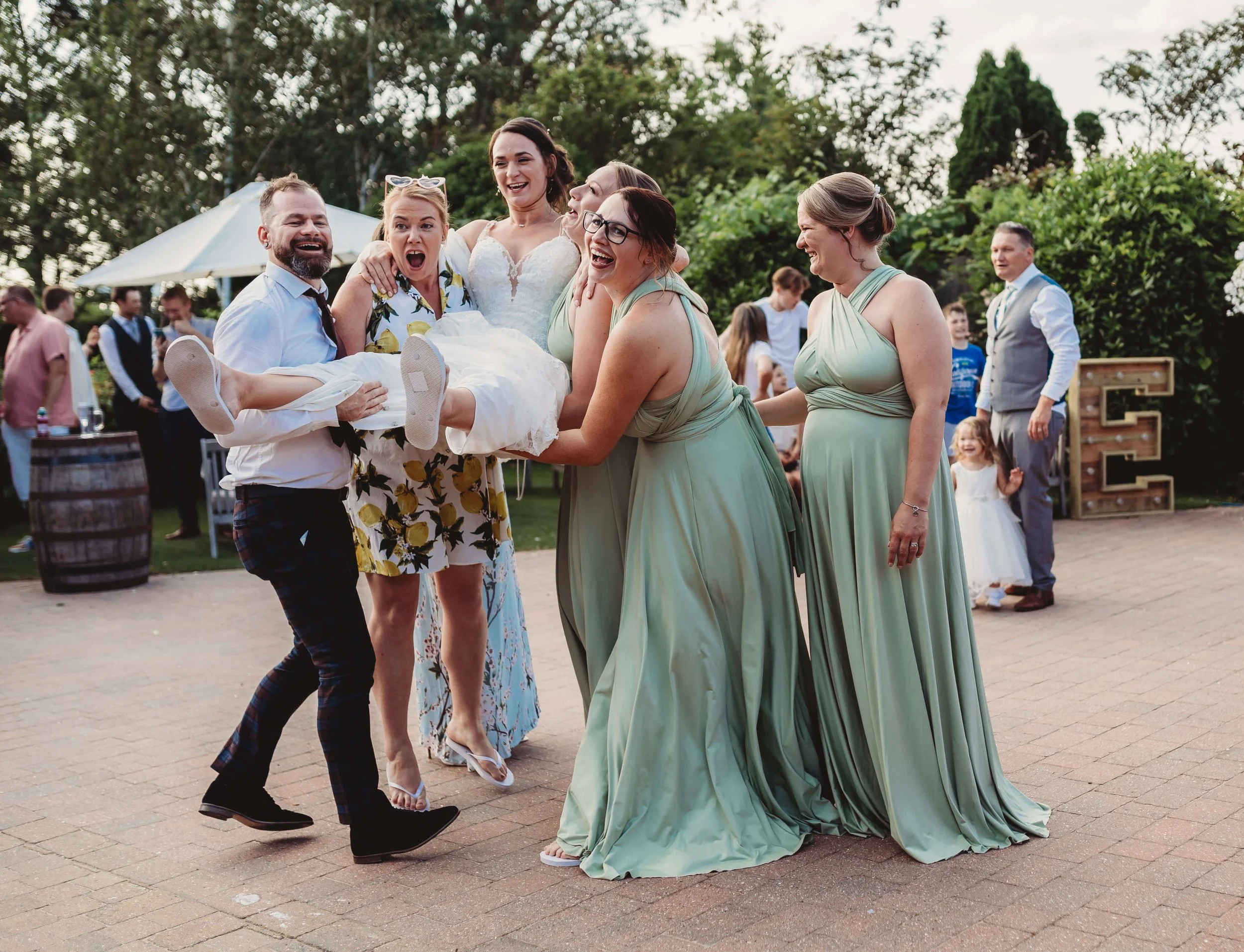 A group of people celebrating at a wedding reception, with a bride being carried by friends, all smiling and laughing outdoors.