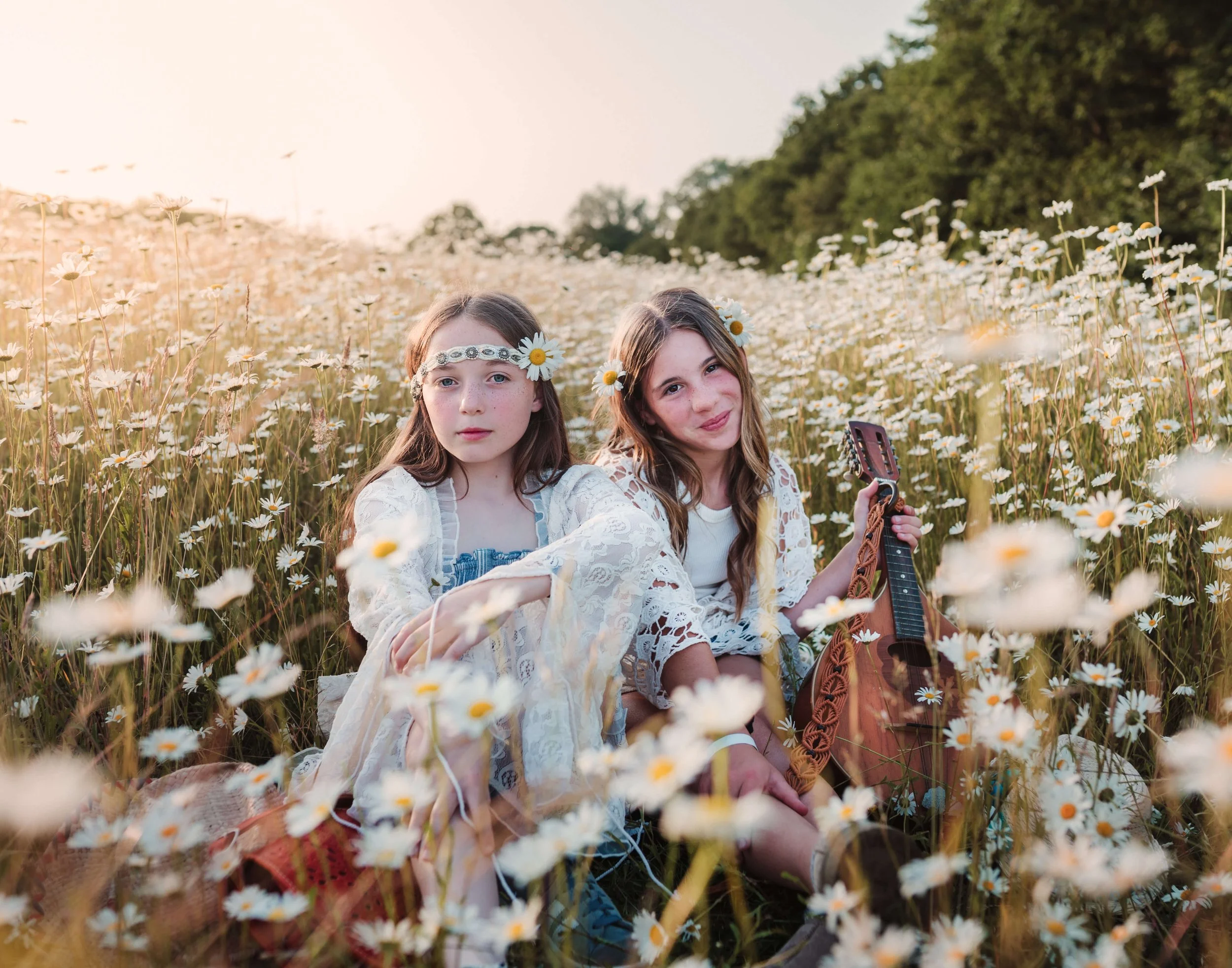 Two young girls sitting in a field of daisies, wearing flower headbands, with one holding a guitar, during sunset.