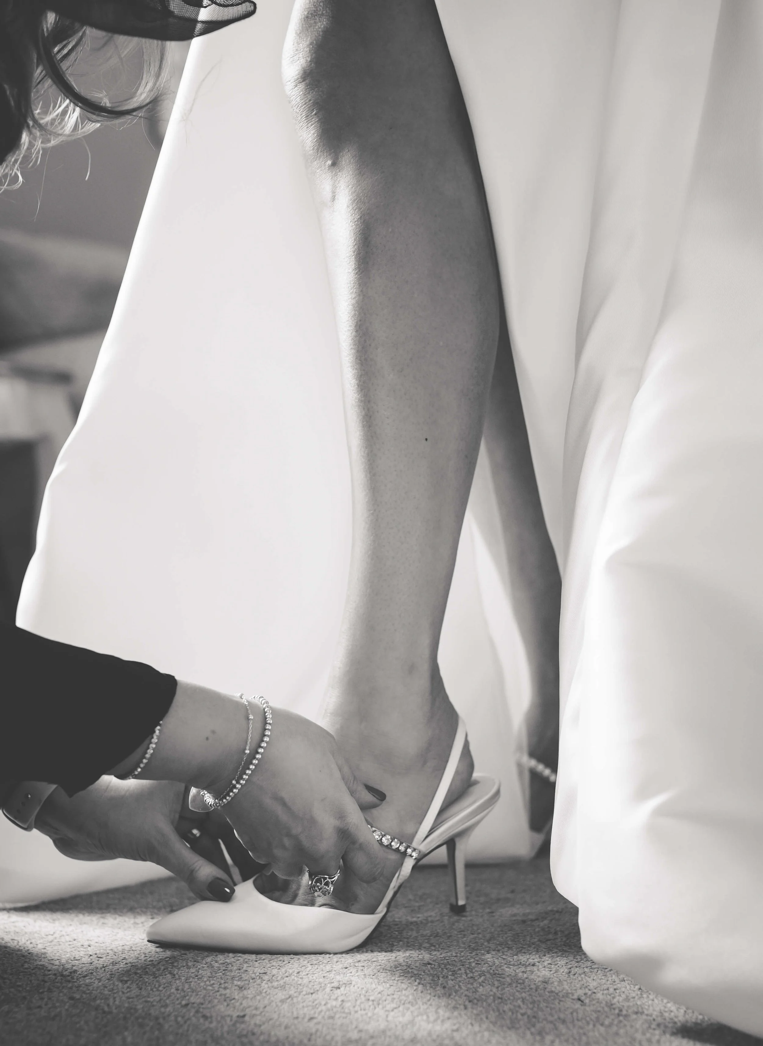A person is helping another person put on a white high-heeled shoe, with only the lower legs and hands visible. The scene appears to involve a fitting or dressing process, possibly for a wedding or special occasion.