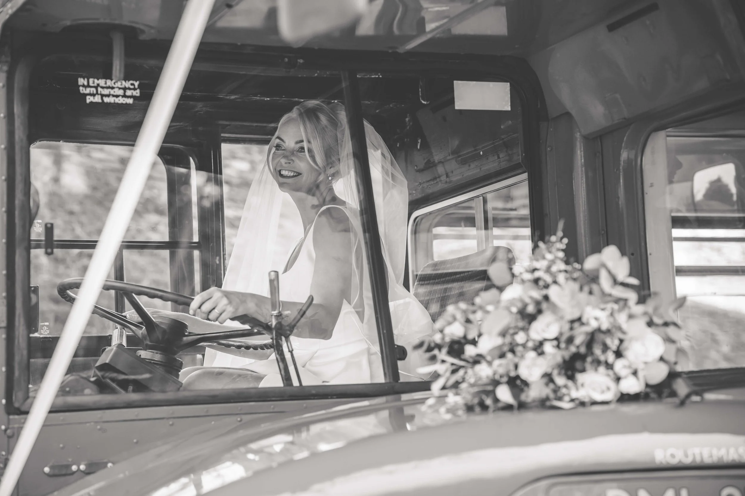 A bride in a wedding dress and veil sitting inside a vintage vehicle with a bouquet of flowers on the dashboard, smiling.