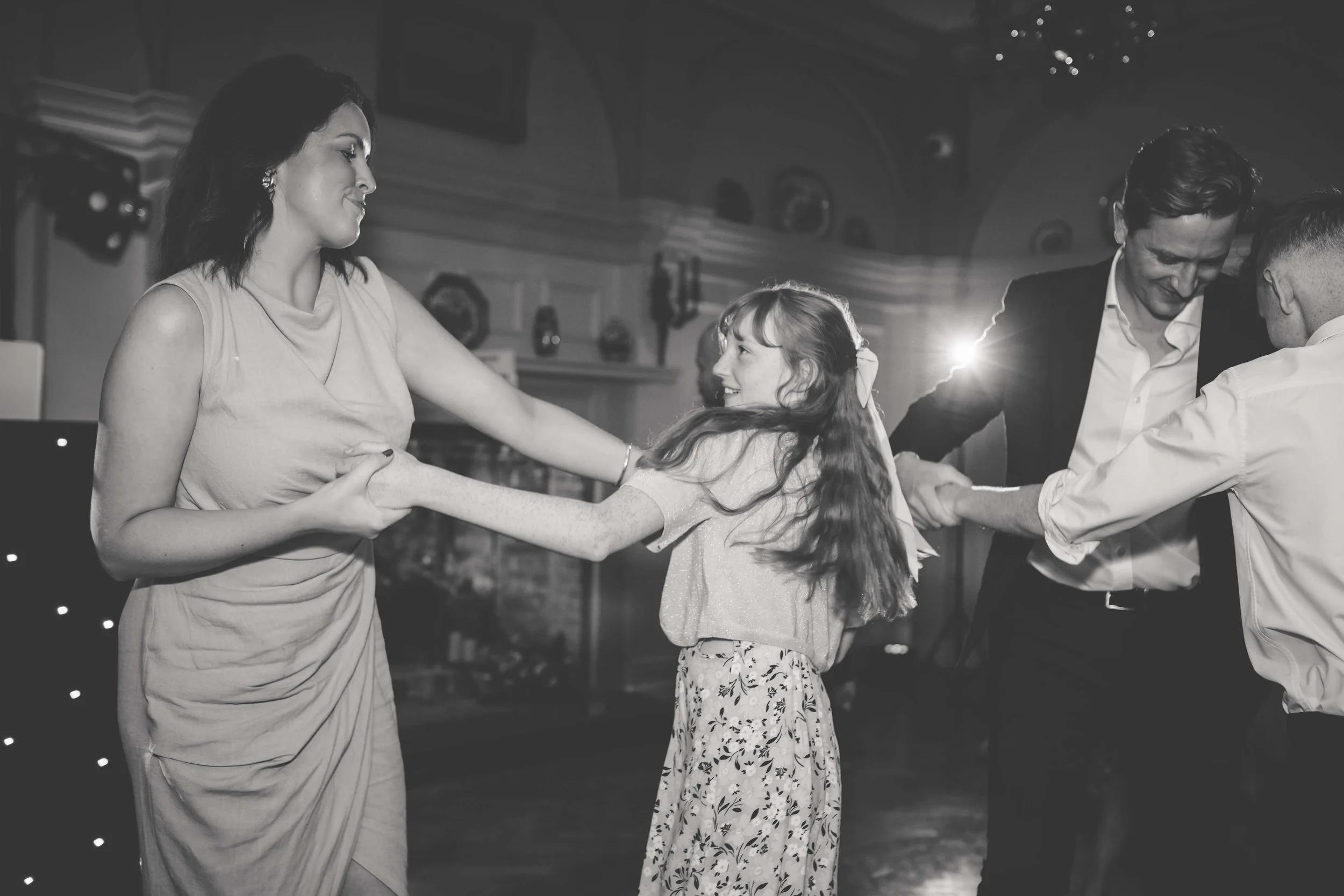A black and white photo of four people dancing at a social gathering. A woman with dark hair wearing a sleeveless dress is holding hands with a young girl with long hair and a floral skirt. Two men, one with dark hair in a suit and another with short