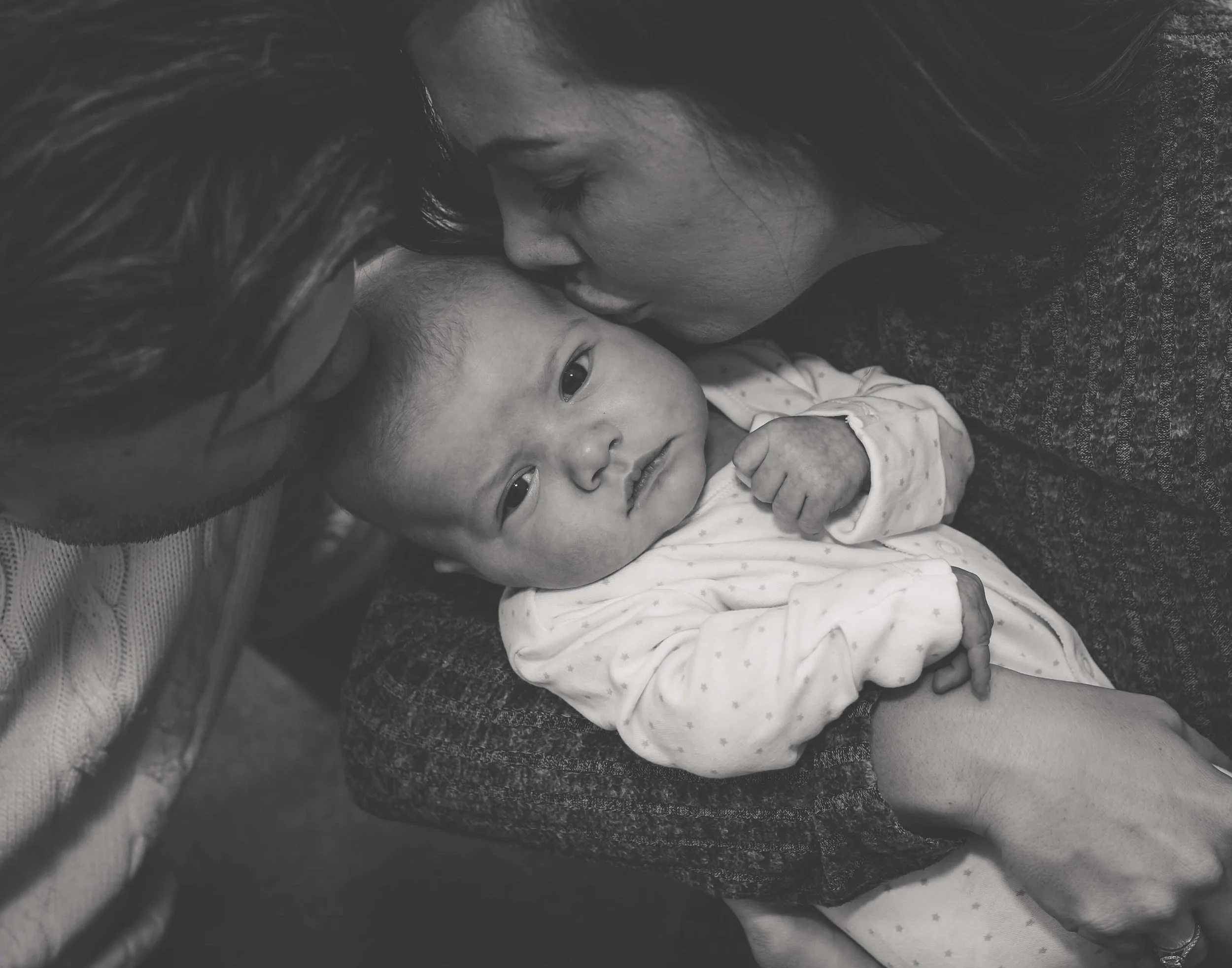 A woman and man leaning in close, kissing a newborn baby on the forehead. The baby is looking at the camera with a calm expression. The woman is resting her head against the baby's head, and the man is close to her.