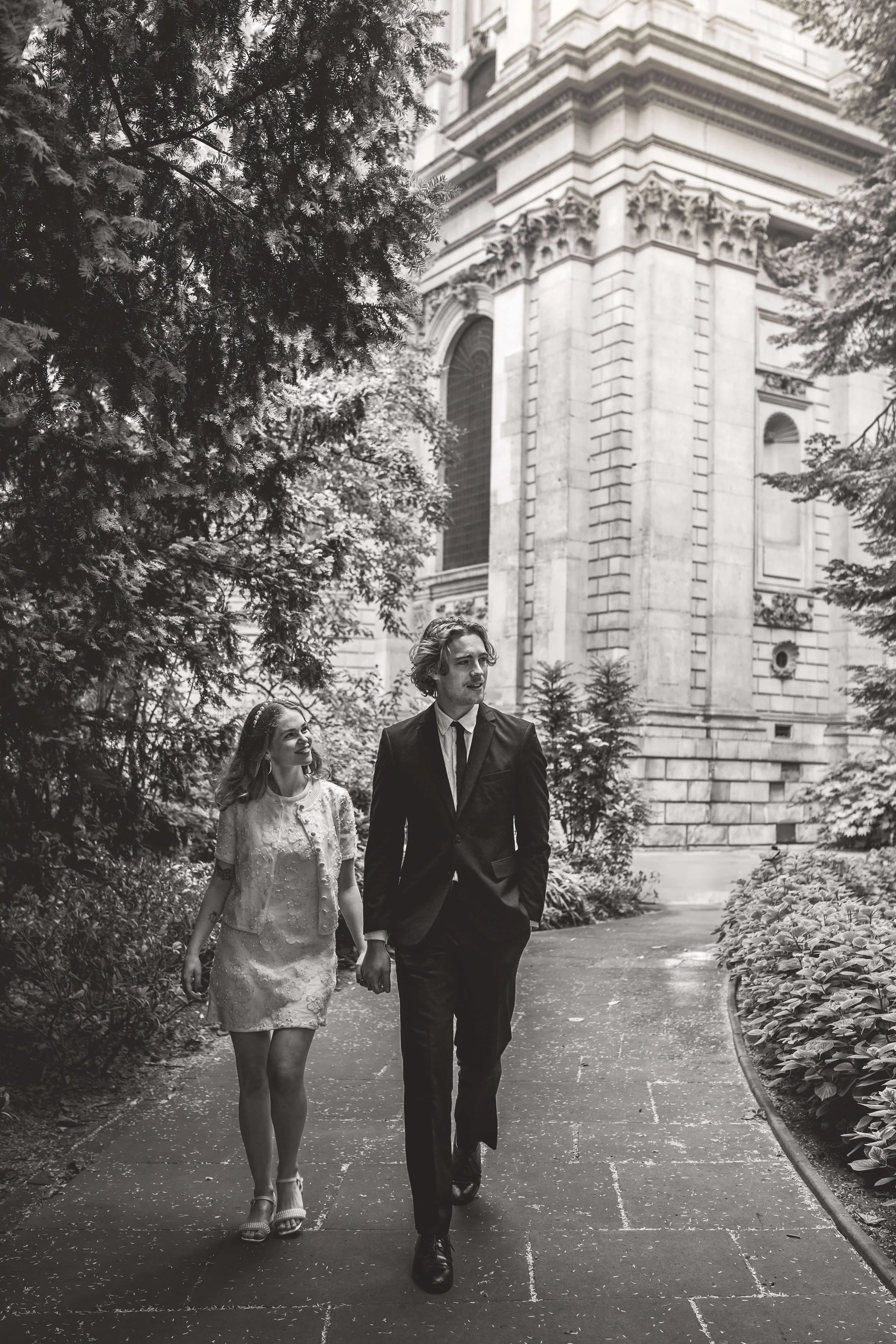 Black and white photo of a couple walking hand in hand along a garden path near a historic building with elaborate stonework and large windows.