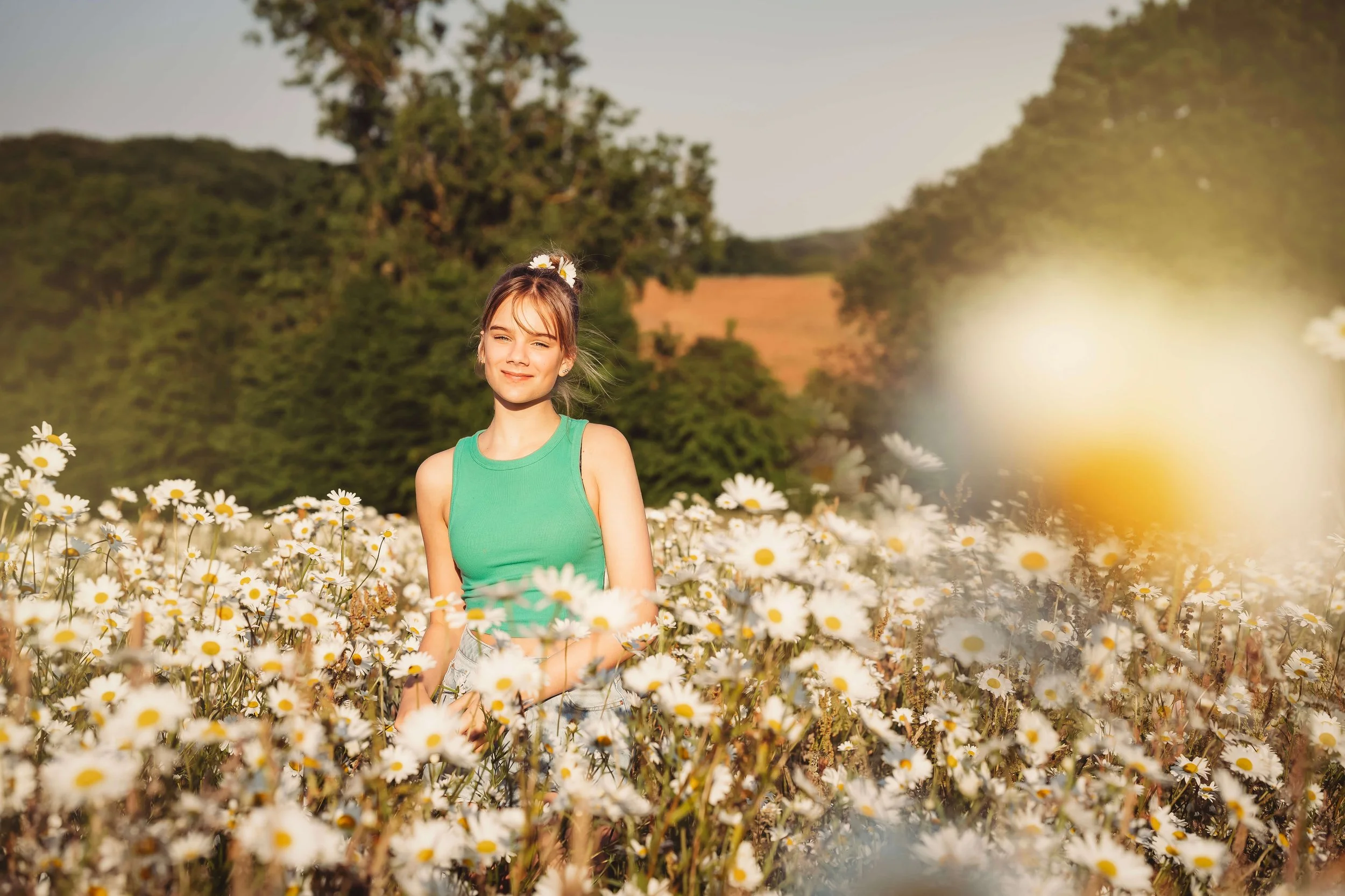 A young woman in a green top standing in a field of daisies during sunset, with trees and hills in the background.