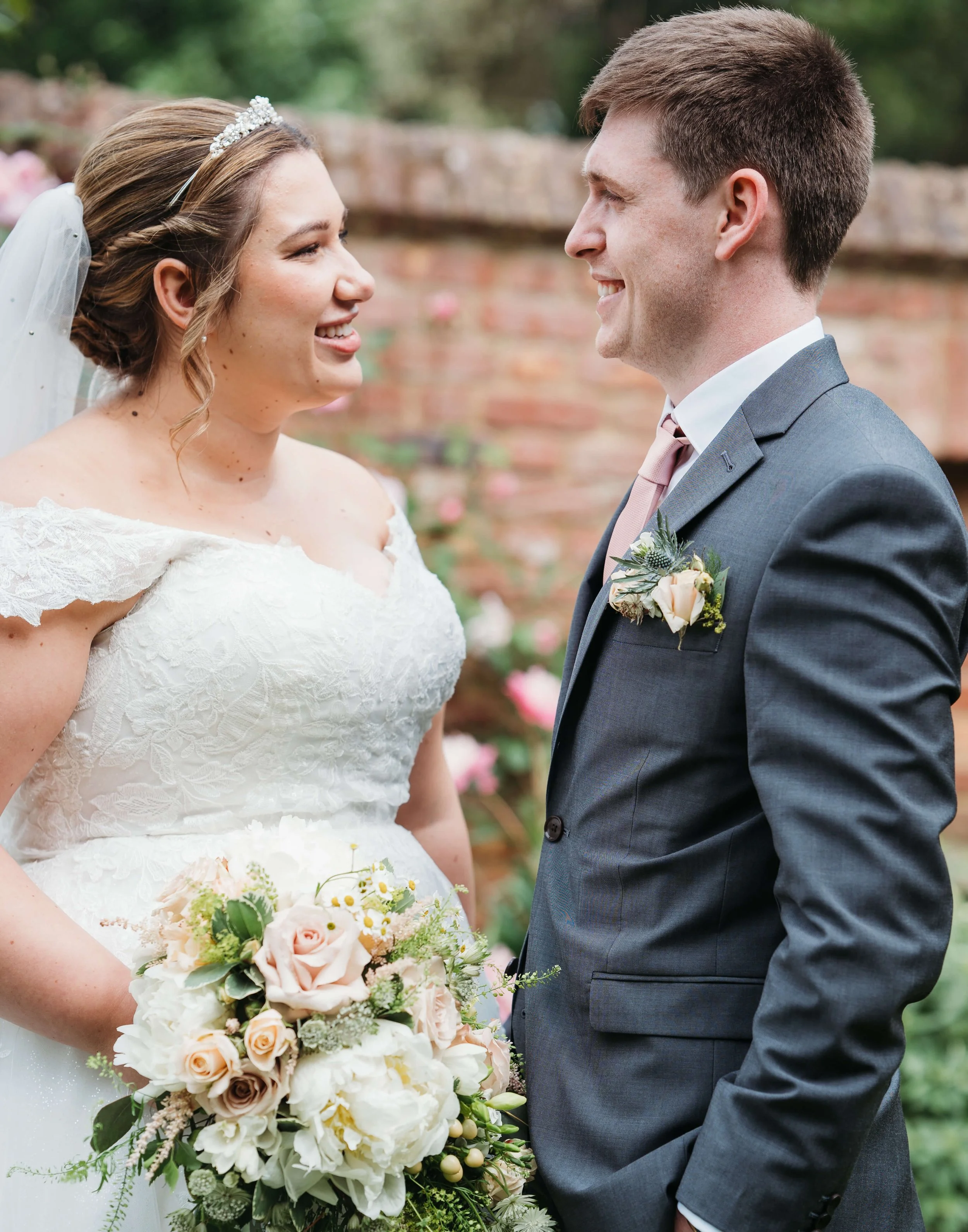 A bride and groom face each other smiling outdoors, with the bride holding a bouquet of flowers and wearing a white wedding dress, while the groom wears a gray suit with a boutonniere.