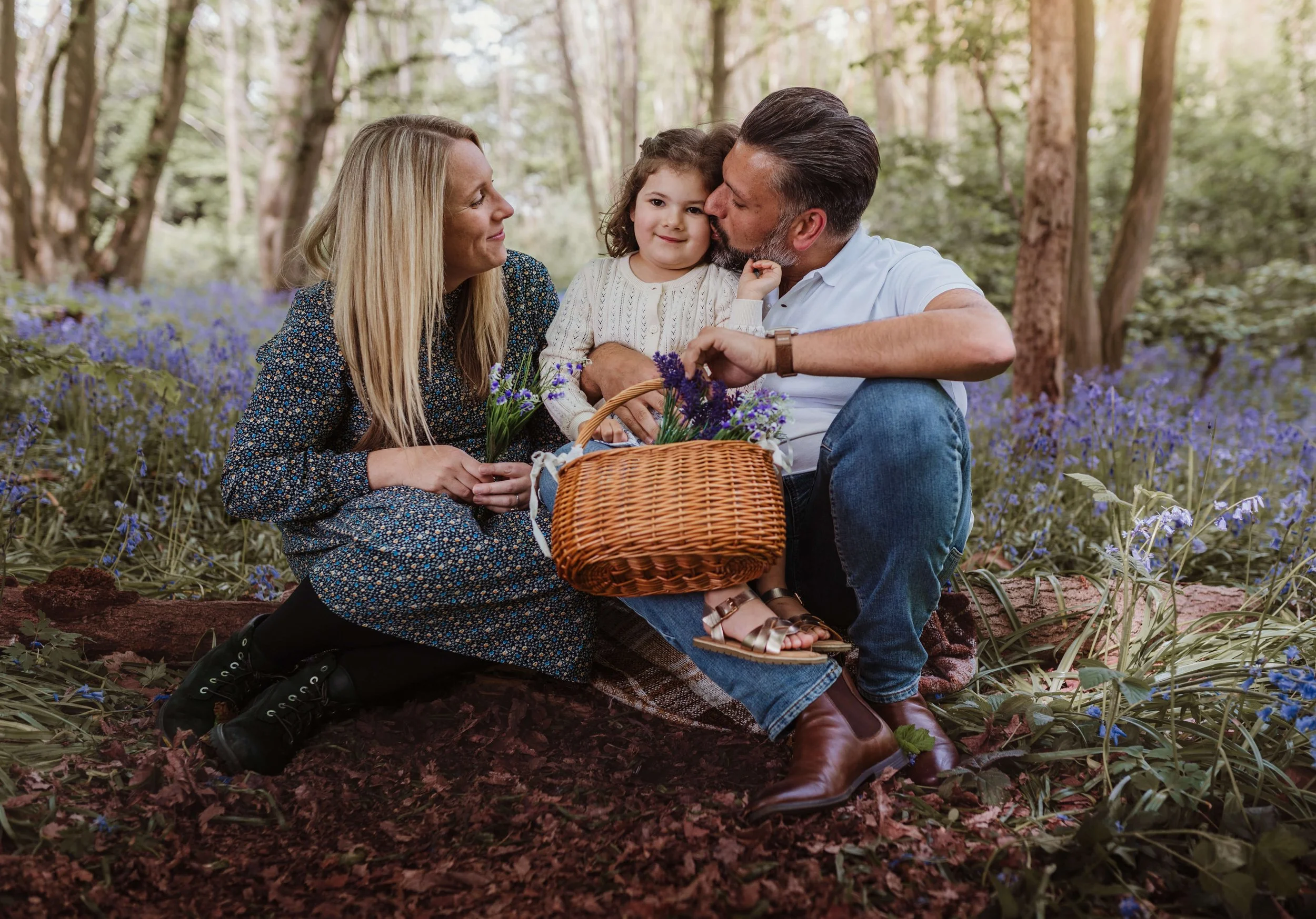 A family of three sitting on the ground in a forested area, having a picnic among blue flowers. The mother, father, and young daughter are closely gathered, with the father kissing the daughter on the cheek. They have a wicker picnic basket with flow