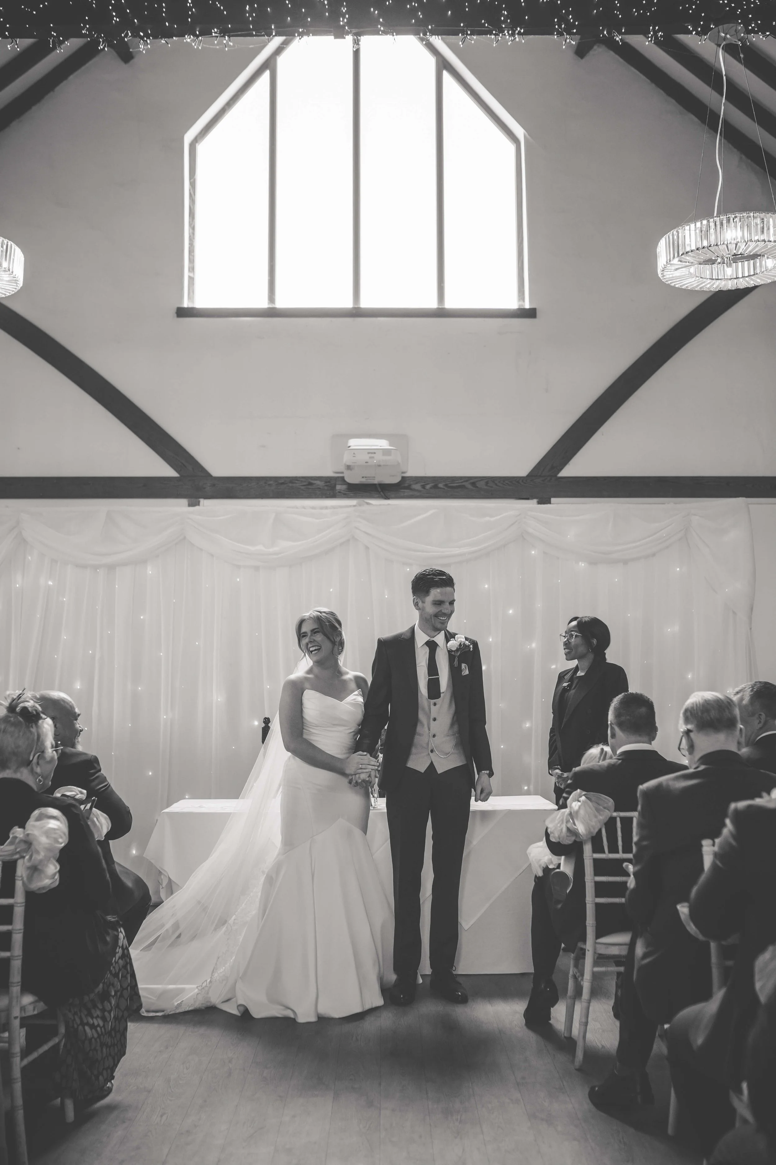 Black and white photo of a wedding ceremony with a bride and groom holding hands and smiling at each other, surrounded by seated guests, with a decorated backdrop of curtains and fairy lights.