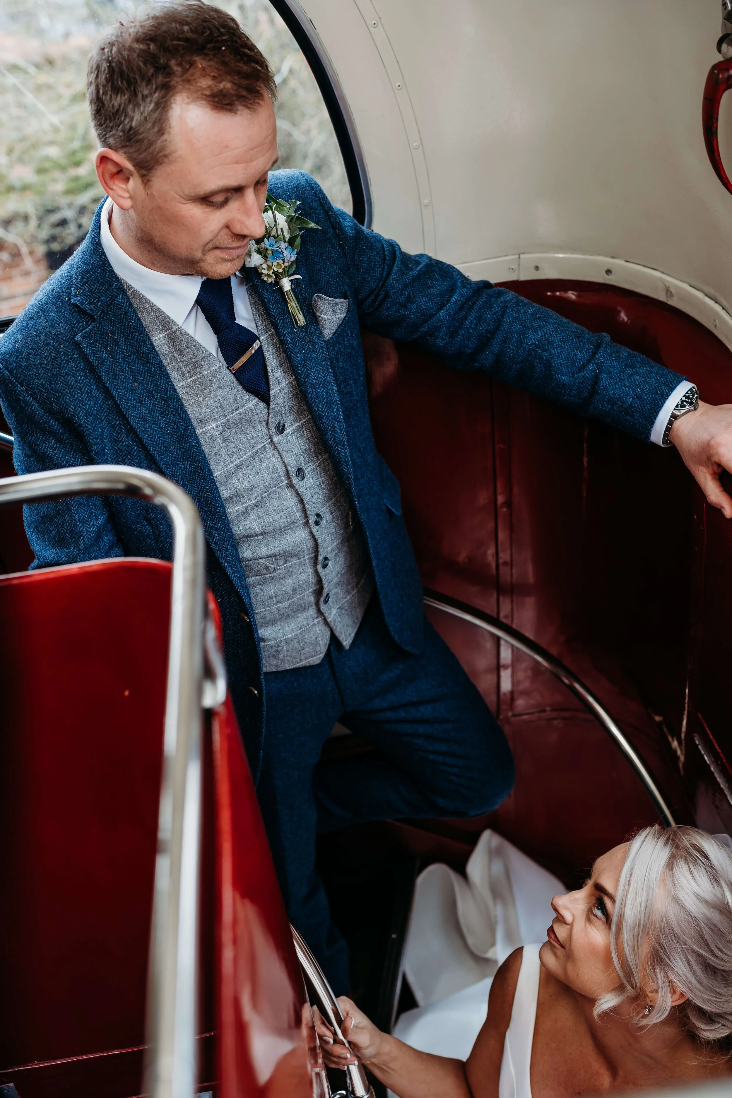A groom dressed in a blue suit with a gray vest and boutonniere is helping a bride with silver hair, who is sitting inside a red vintage bus. The bride is looking up at the groom.