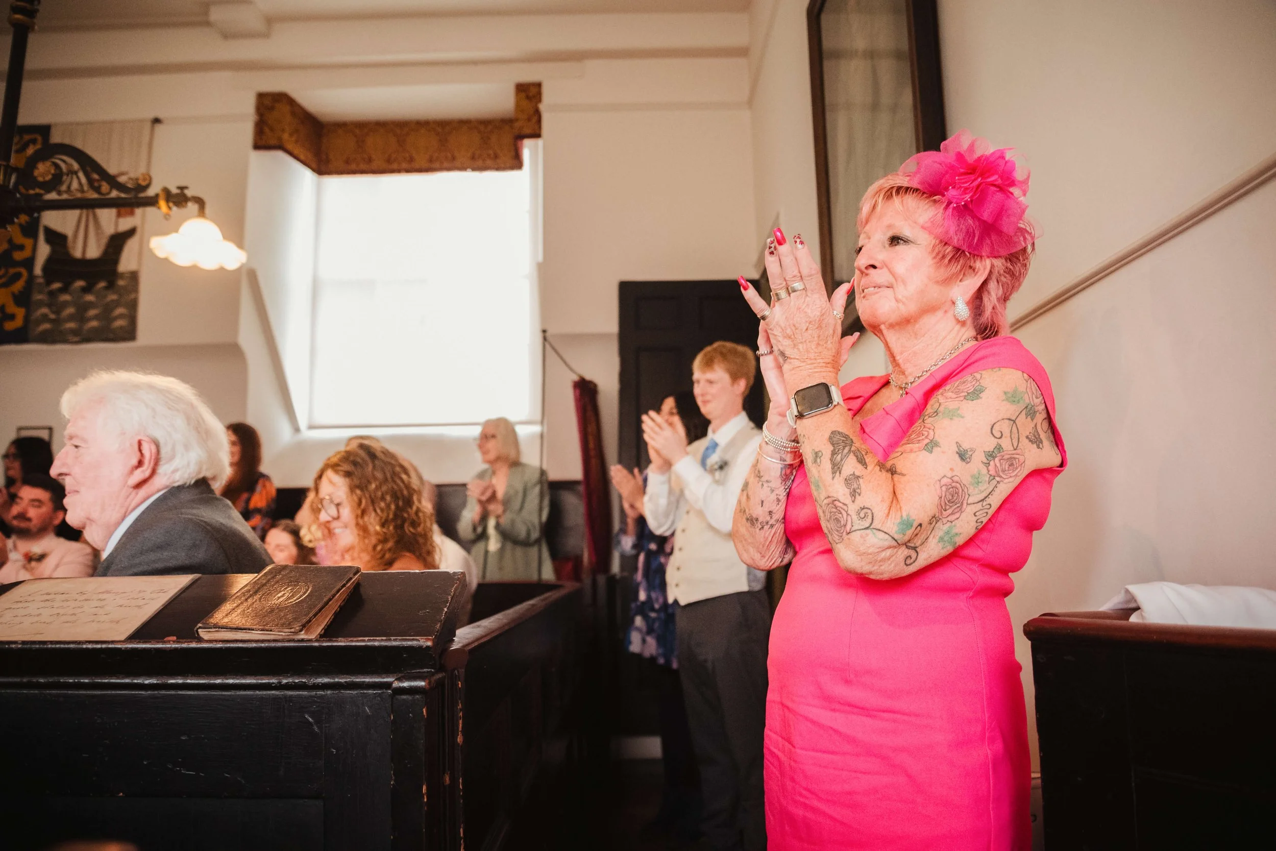 An elderly woman with pink hair and floral tattoos on her arms in a pink dress clapping with her hands near her face, at a formal event or gathering in a church or hall.