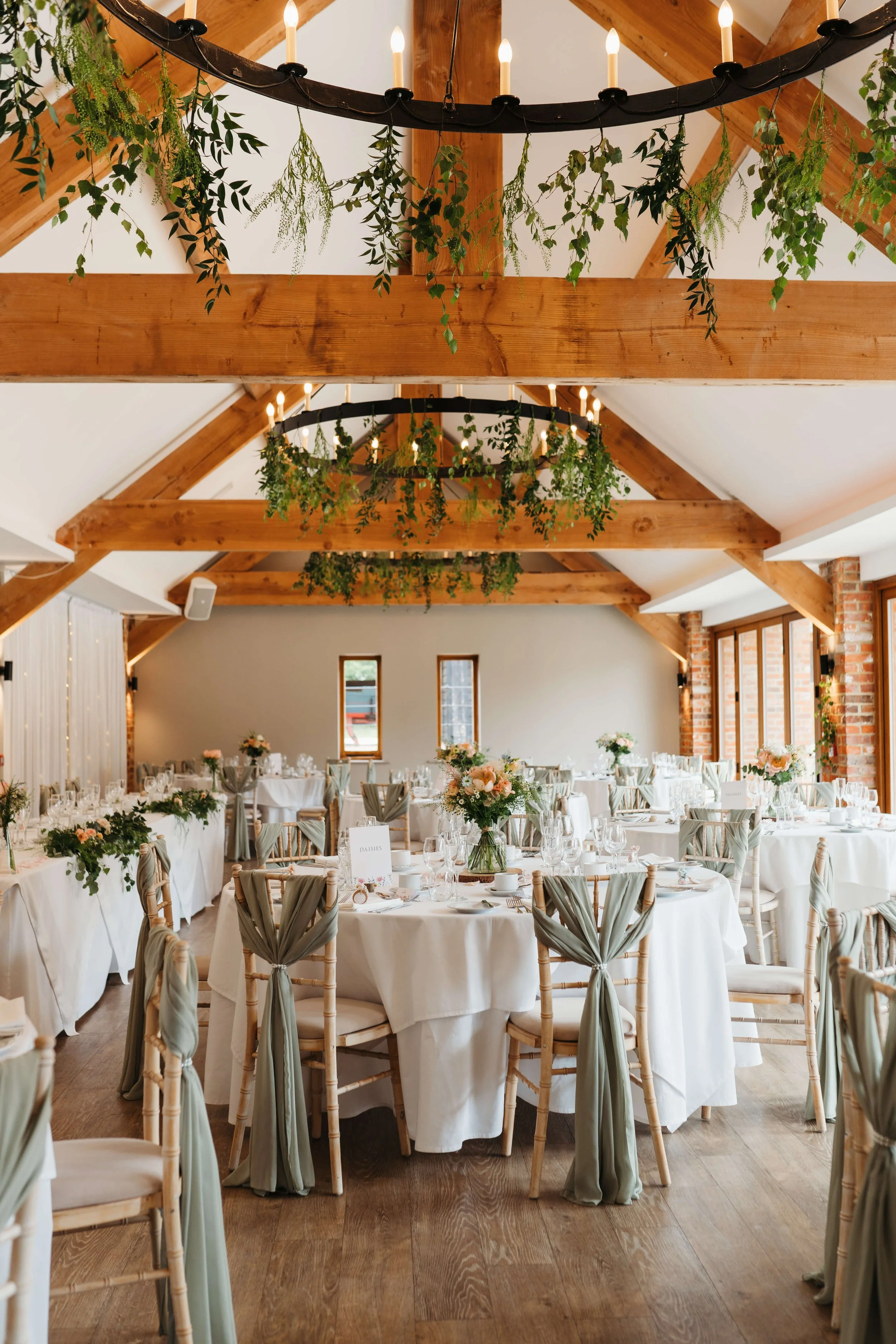A decorated banquet hall with round tables covered in white tablecloths, adorned with floral centerpieces, set for a formal event. The ceiling has wooden beams and features hanging greenery and two circular black chandeliers with lit candles.