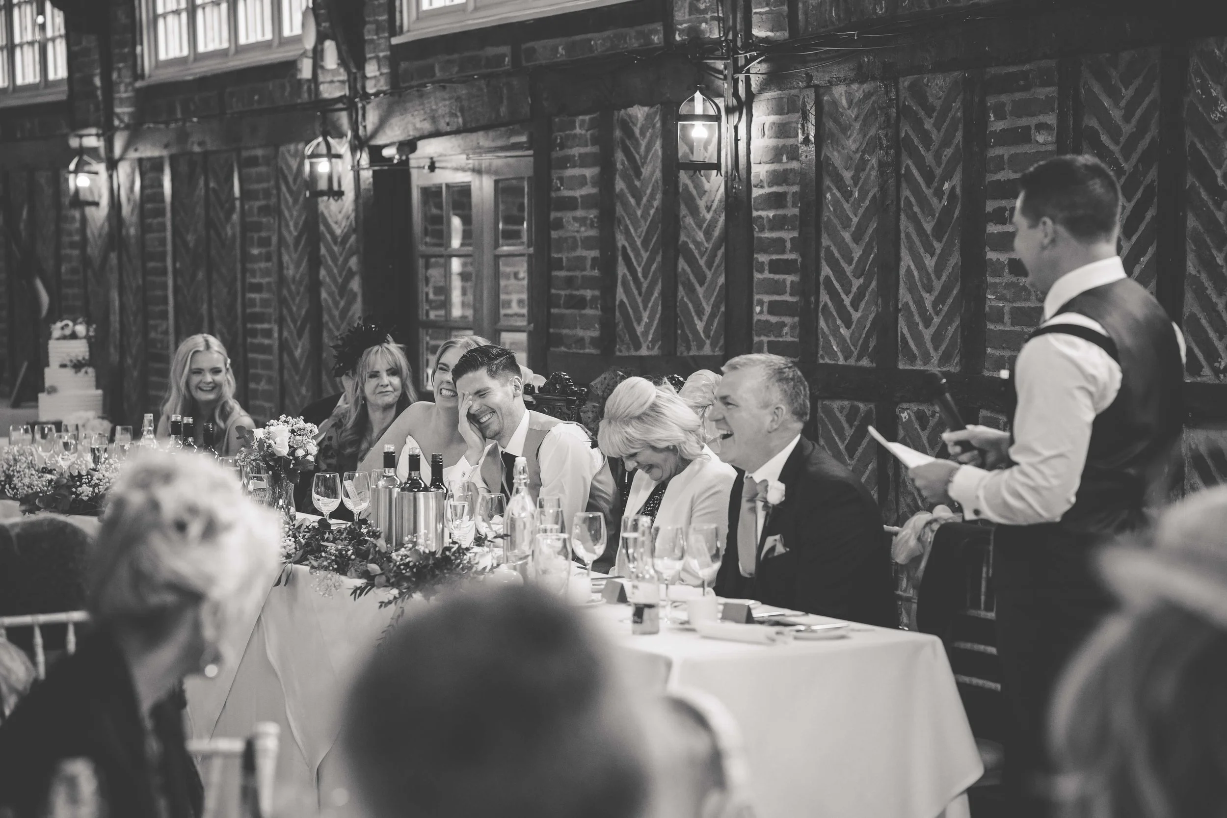 People seated at a dining table during a wedding reception, sharing a laugh, with a man giving a toast, in a rustic brick-walled venue.