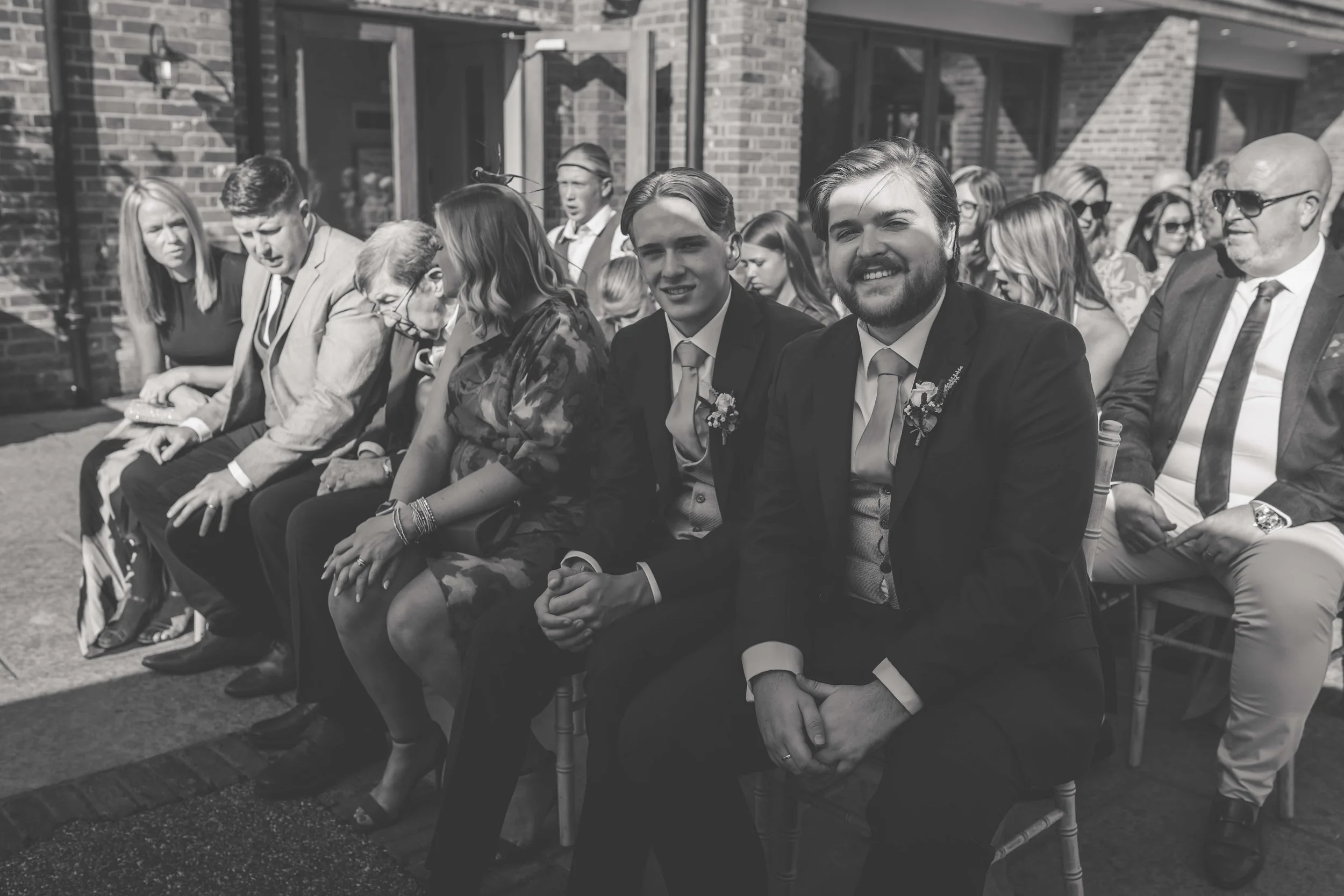 Black and white photo of wedding guests sitting outdoors, with two men in suits smiling at the camera in the front row. The background shows a brick building and a crowd of seated guests.