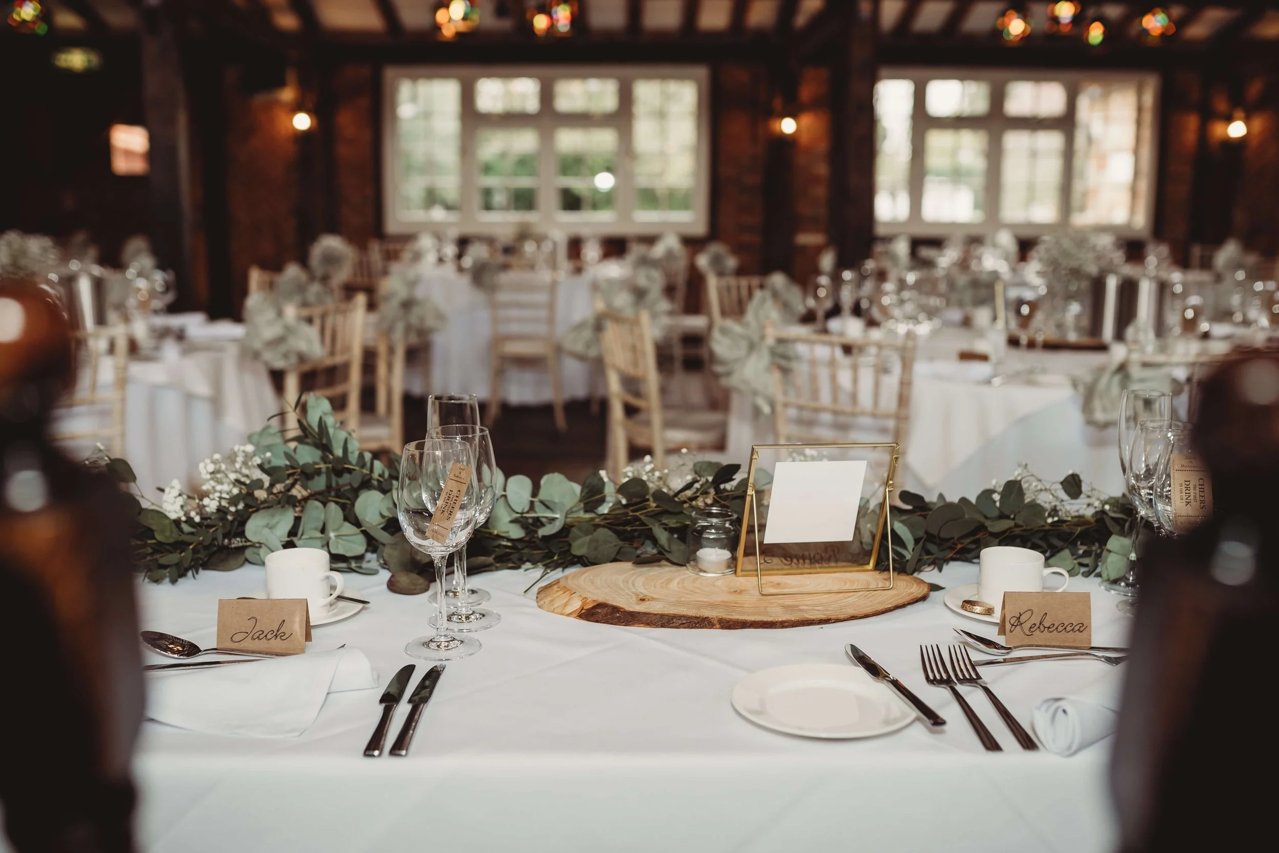 Wedding reception table setup with white tablecloth, green foliage centerpiece on a wooden slab, place cards labeled 'Jack' and 'Rebecca', wine glasses, white coffee mug, silverware, a picture frame, and small candles in a rustic venue with wooden wa