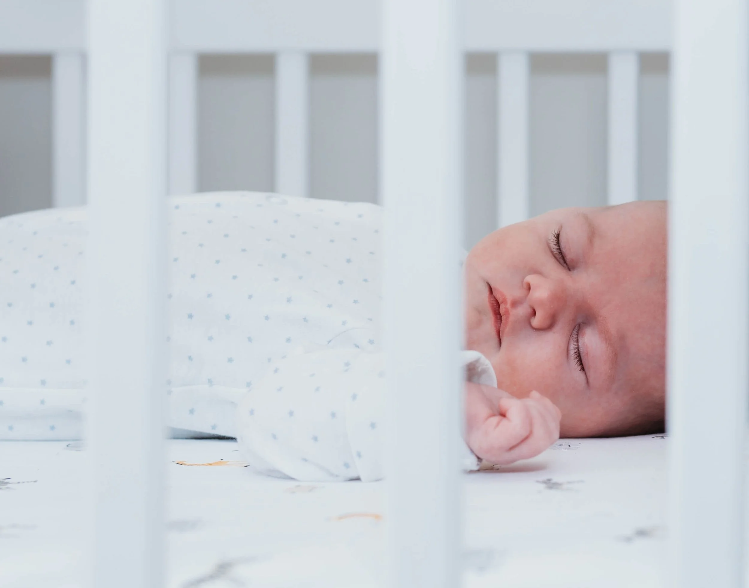 A sleeping baby lying on a white crib mattress, seen through the crib's bars.