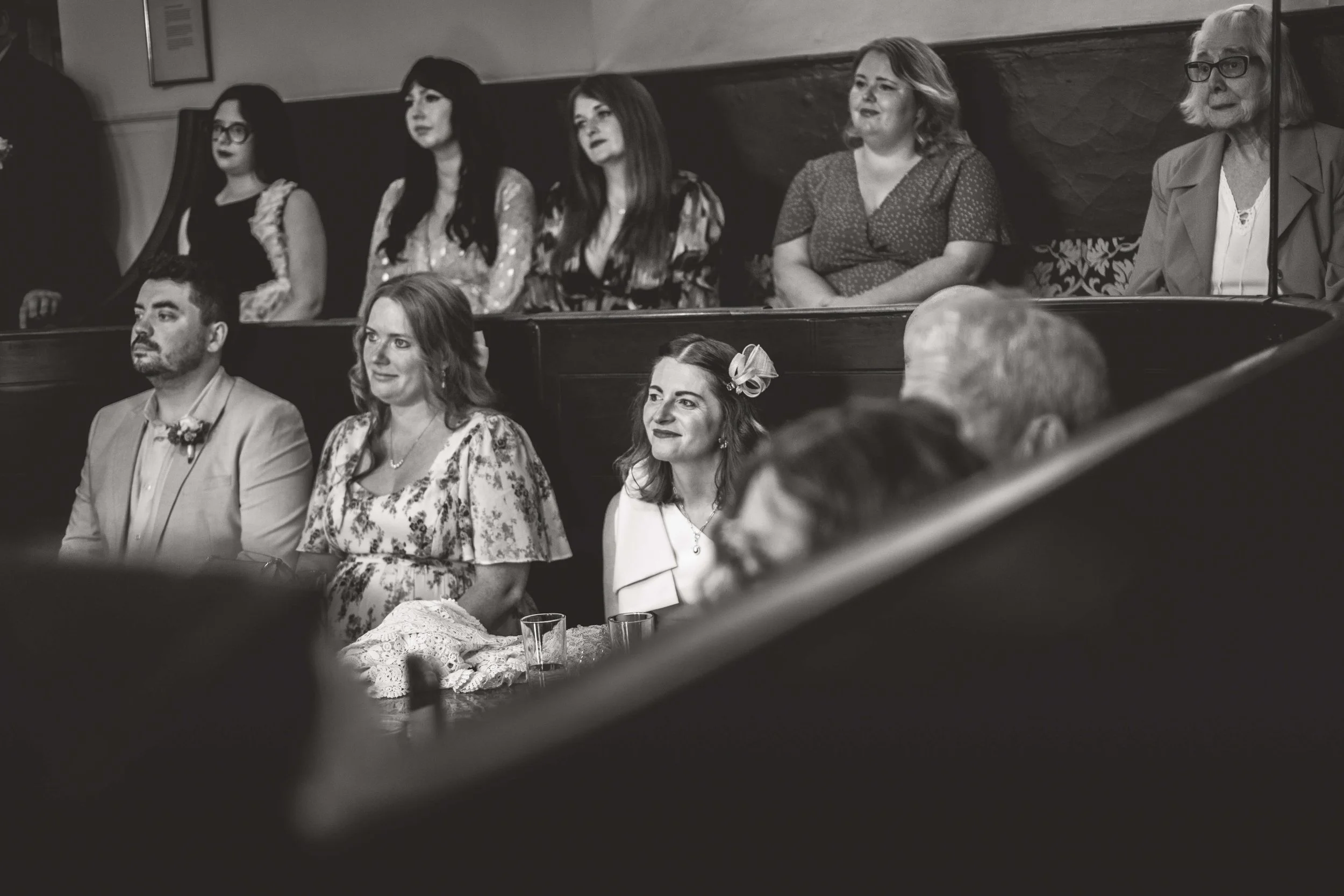 Black and white photo of people seated in a church pew during a wedding, including women in dresses with floral accessories, a man in a suit with a boutonniere, and older women.