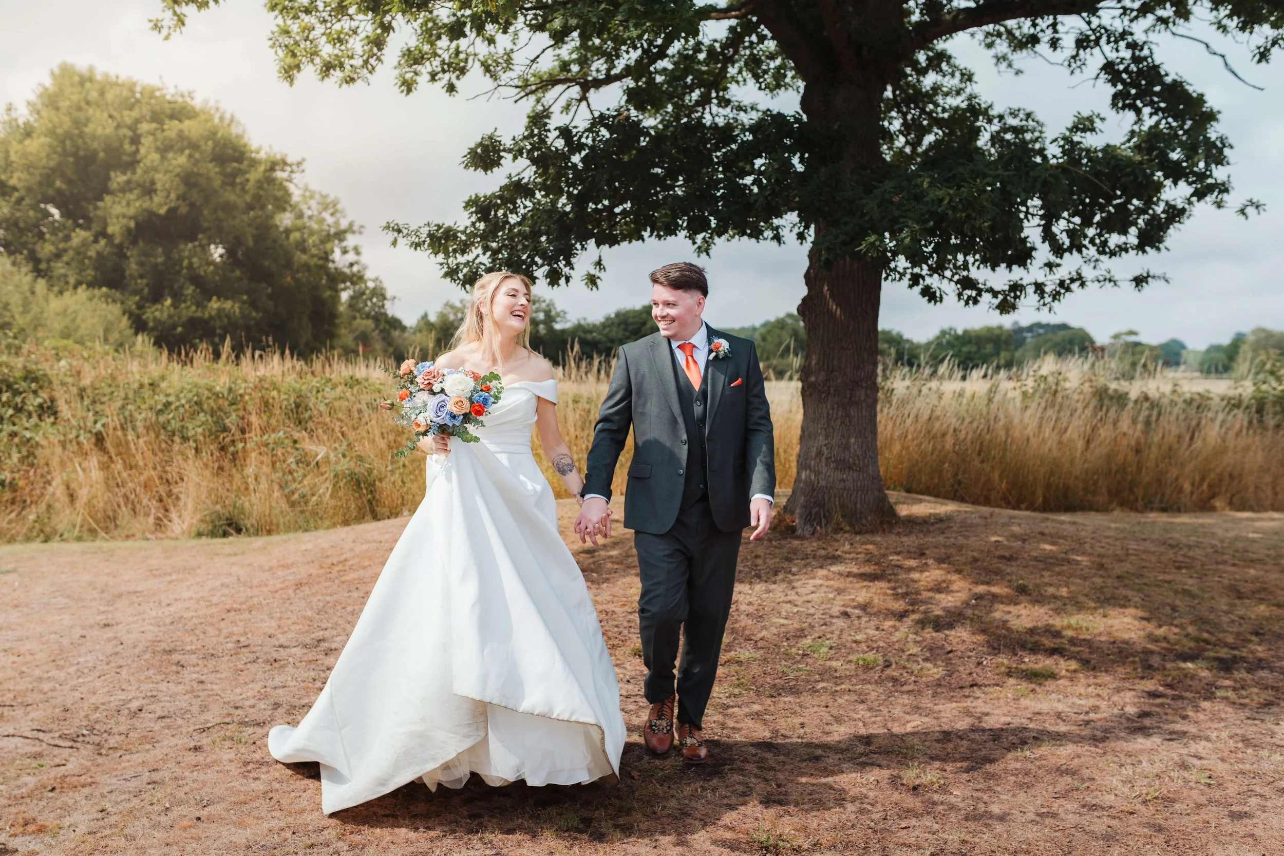 A newlywed couple walking hand-in-hand outdoors, the bride in a white wedding gown holding a bouquet, and the groom in a dark suit with an orange tie, smiling at each other under a large tree with a rural landscape background.