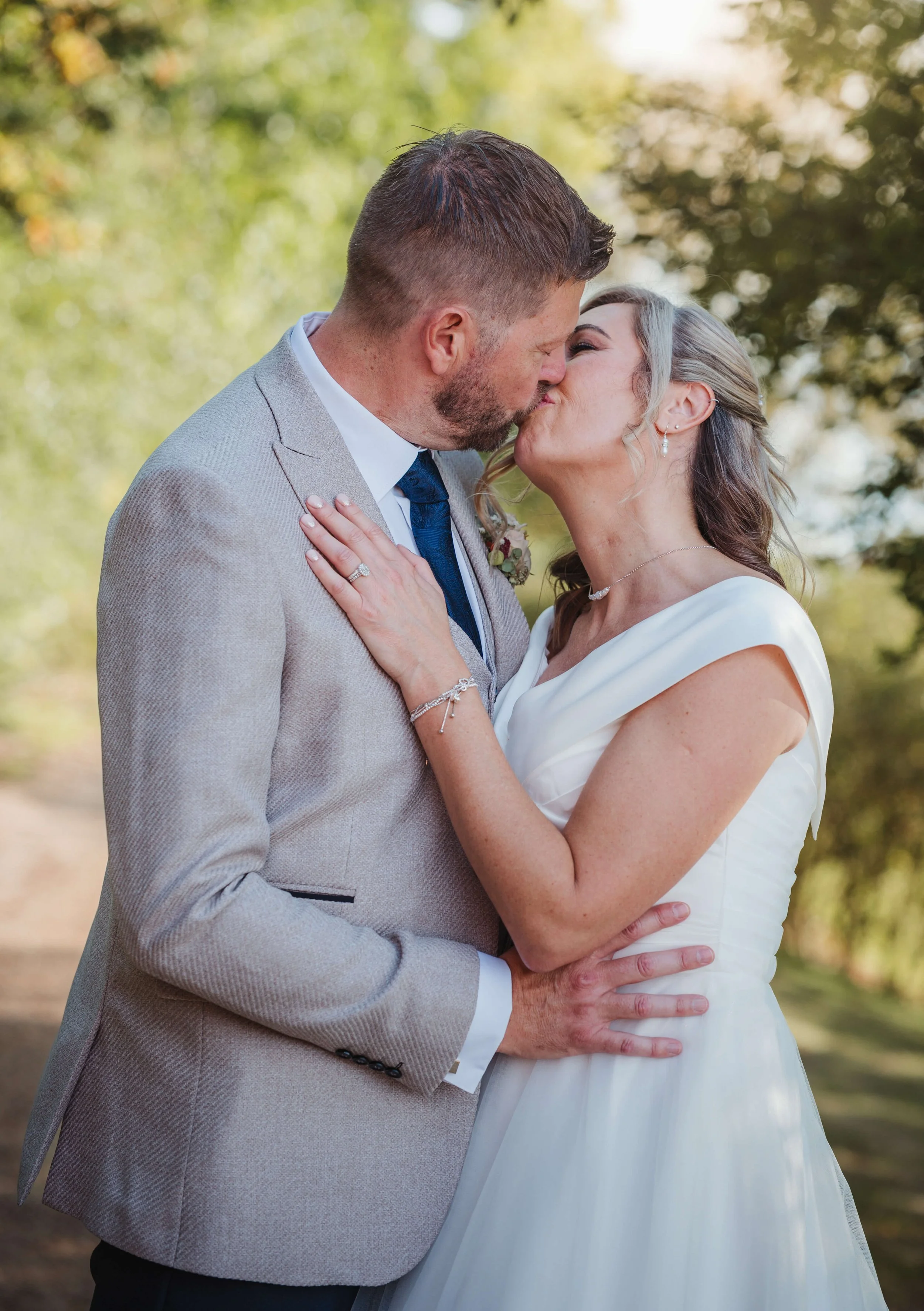 A couple kissing outdoors during a wedding, with the man wearing a light gray suit and the woman in a white wedding dress, surrounded by green trees.