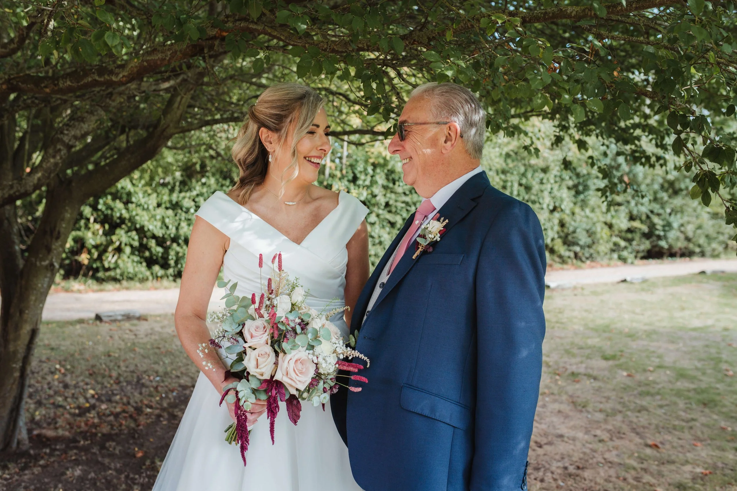 A bride in a white wedding dress holding a bouquet of pink and white roses and greenery, smiling at an older man in a blue suit, pink tie, and glasses, standing outdoors under a tree with green leaves.