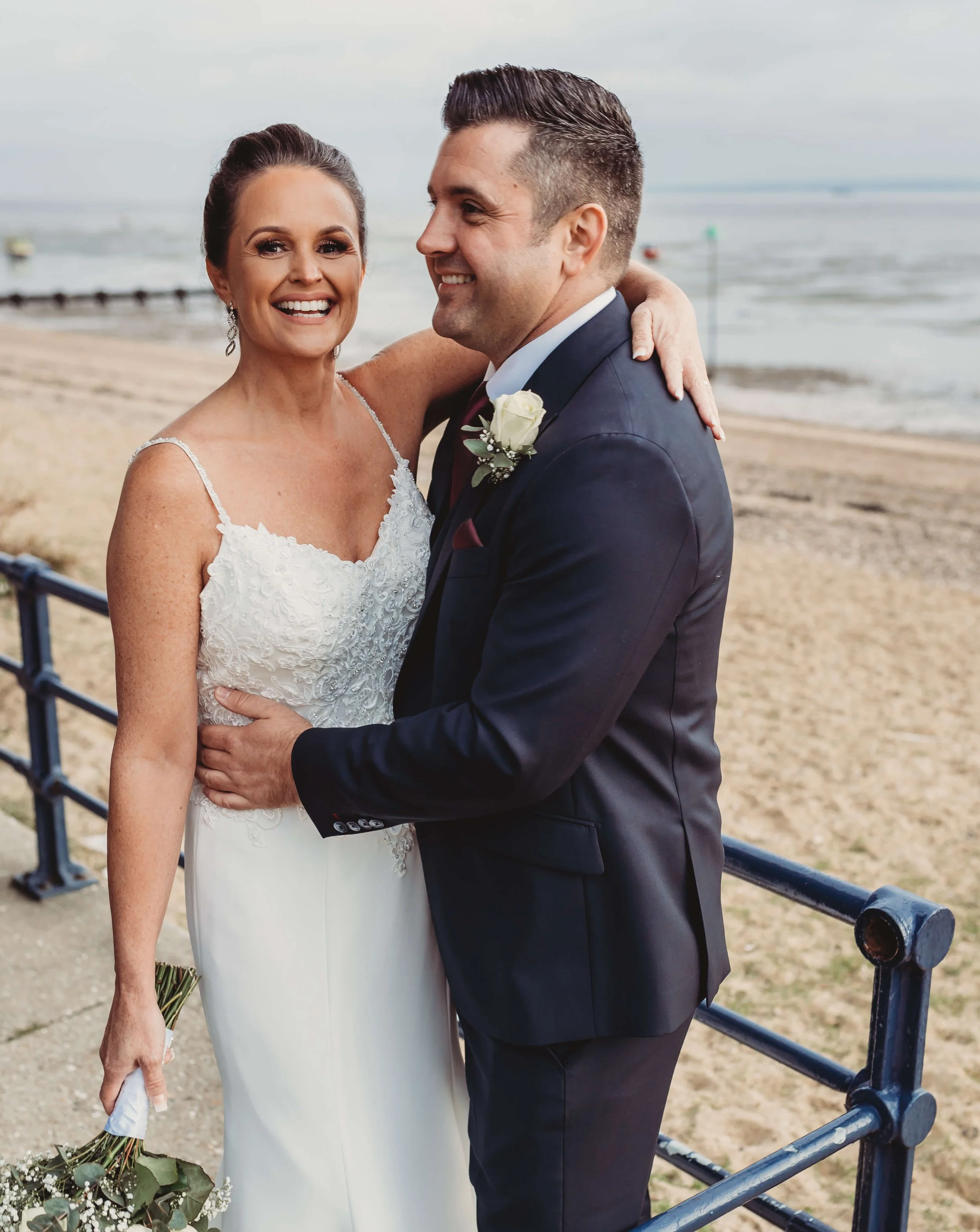 A bride and groom stand close together on a beach, smiling, with the bride holding a small bouquet and the groom wearing a boutonniere, as a railing separates them from the sandy shore and water in the background.
