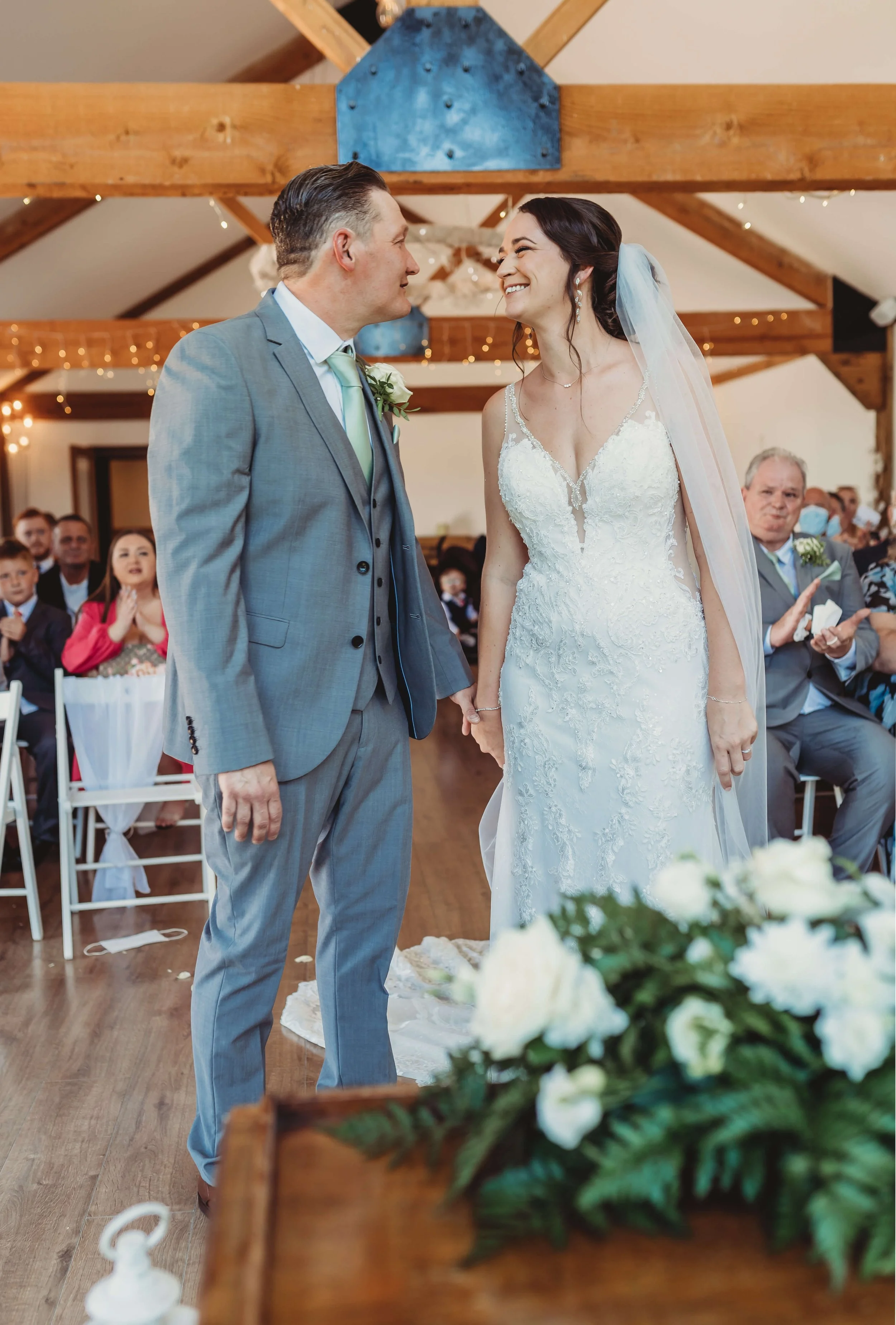 A bride and groom holding hands and smiling at each other during their wedding ceremony inside a decorated venue with wooden beams and fairy lights. Guests are seated in the background, some clapping and watching the couple.