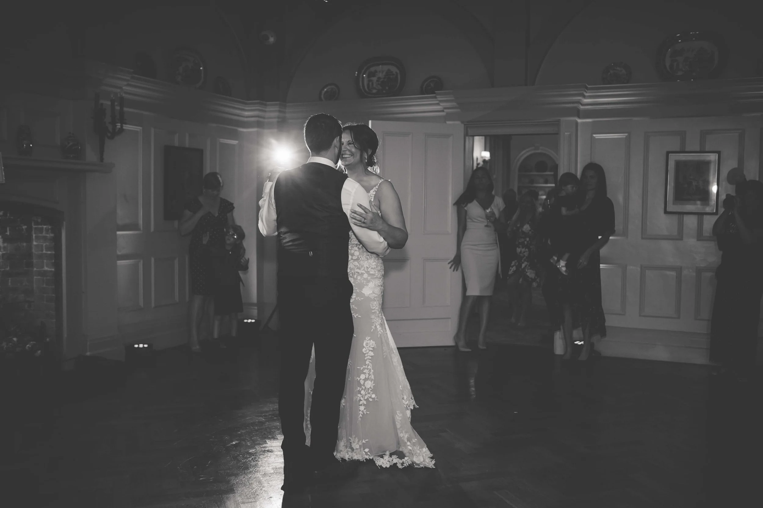 A couple is dancing at their wedding reception, with the bride in a lace gown and the groom in a vest and shirt, surrounded by wedding guests in an elegant room.