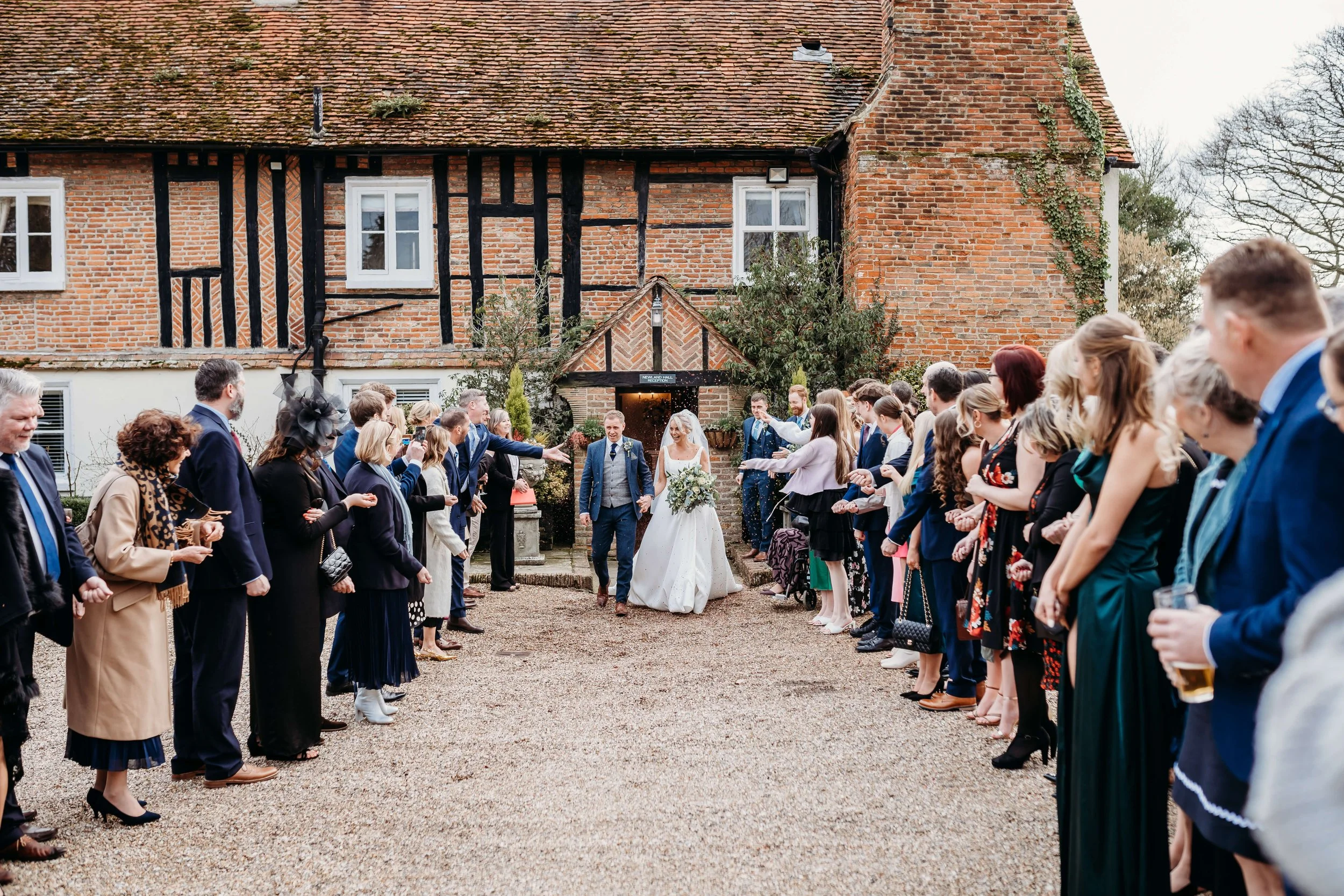 Bride and groom walking out of a brick building with black timber framing, surrounded by friends and family cheering and throwing confetti.