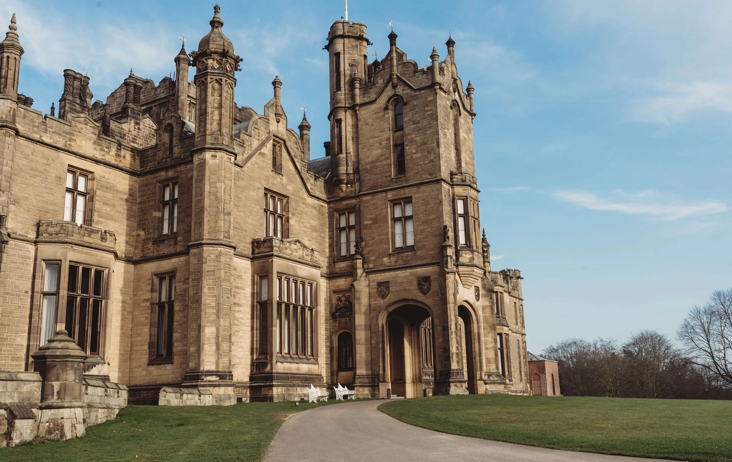 A historic castle with tall towers and stone walls, surrounded by a well-maintained lawn and a curved pathway leading to the entrance, under a clear blue sky.