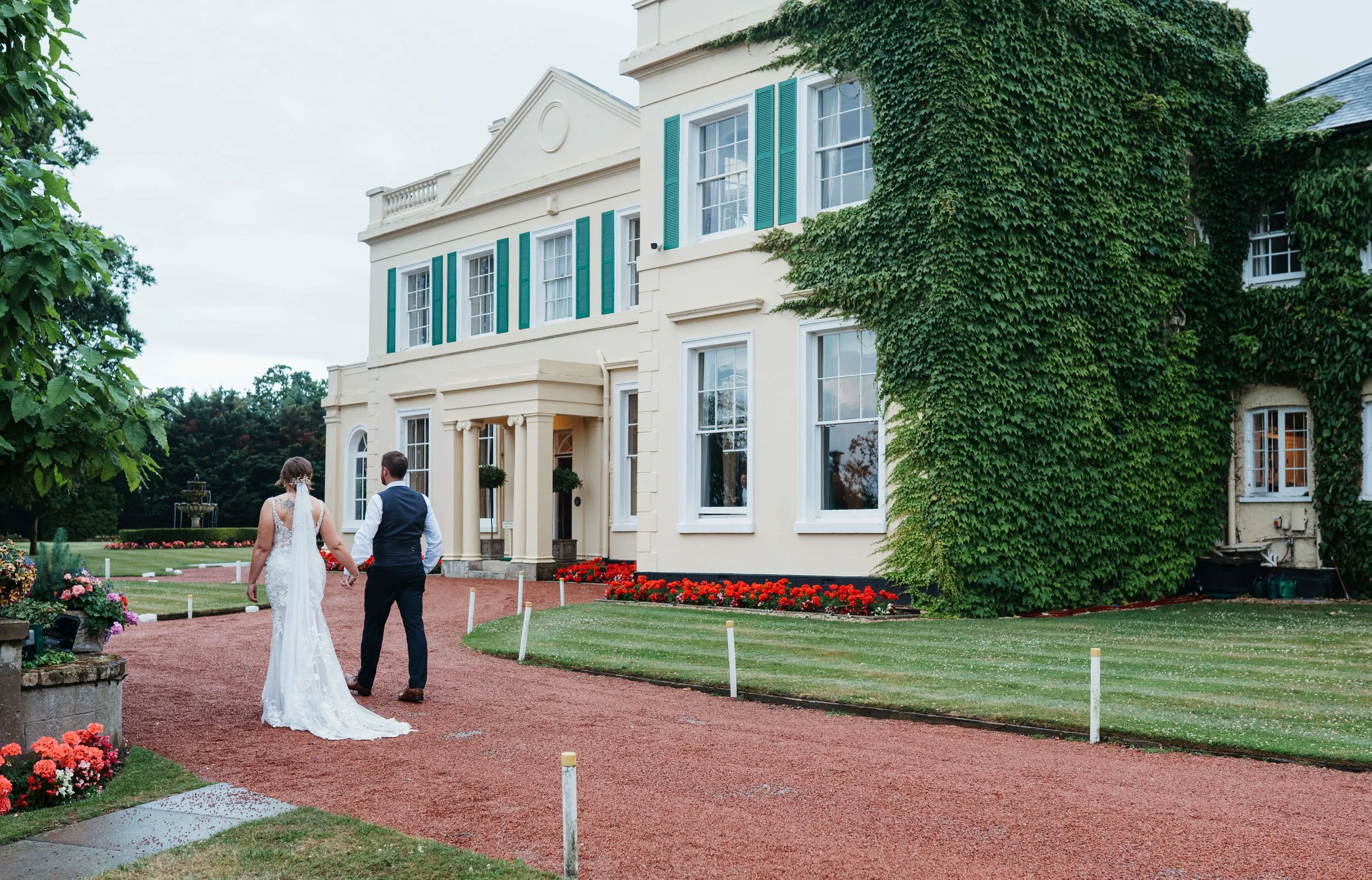 A bride and groom walking on a gravel path outside a large, elegant white building with green shutters and ivy growing on the wall, surrounded by manicured lawns and flower beds.