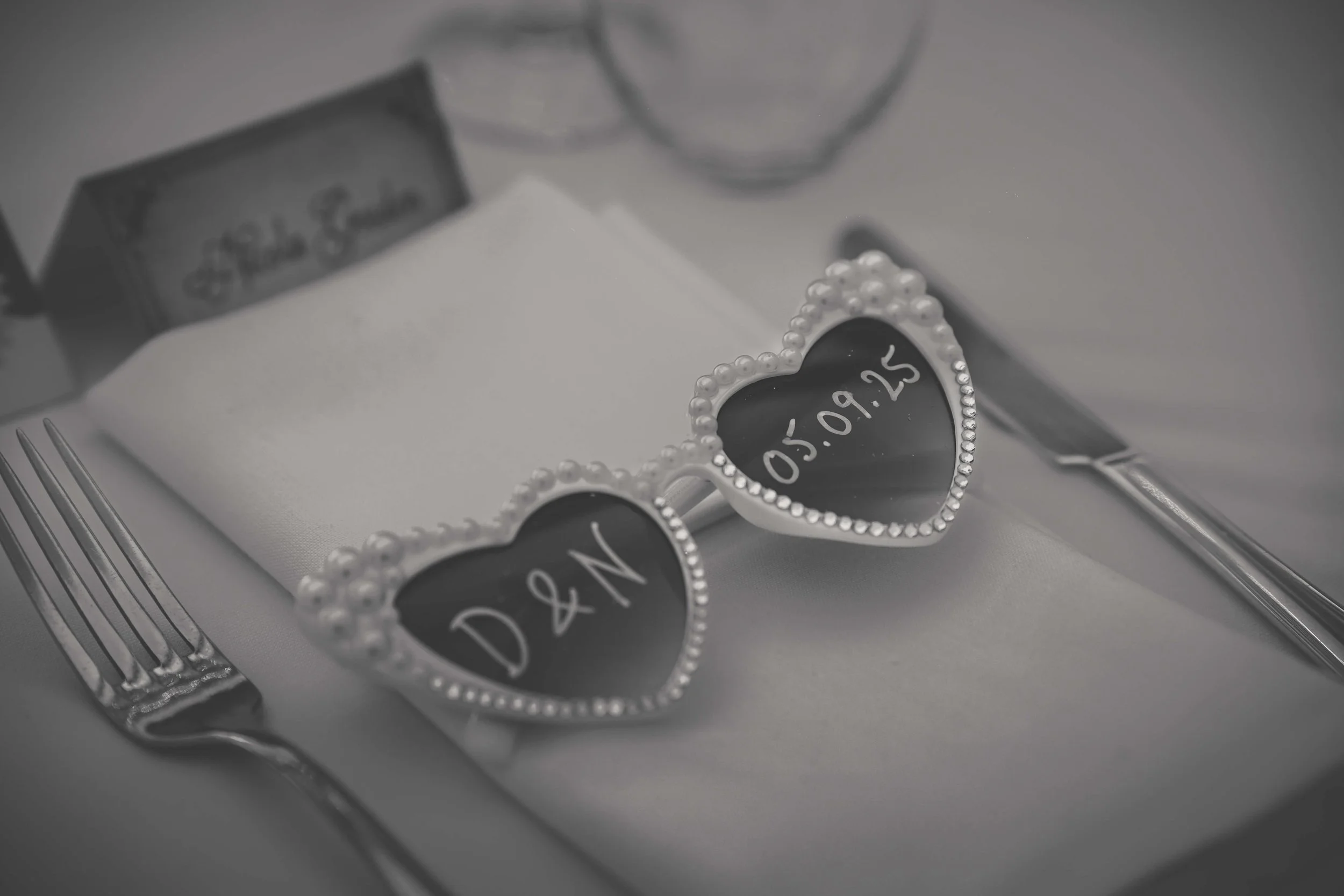Heart-shaped glasses with the date 05.09.25 written on the lenses, resting on a folded napkin on a place setting with a fork and knife, blurred background.