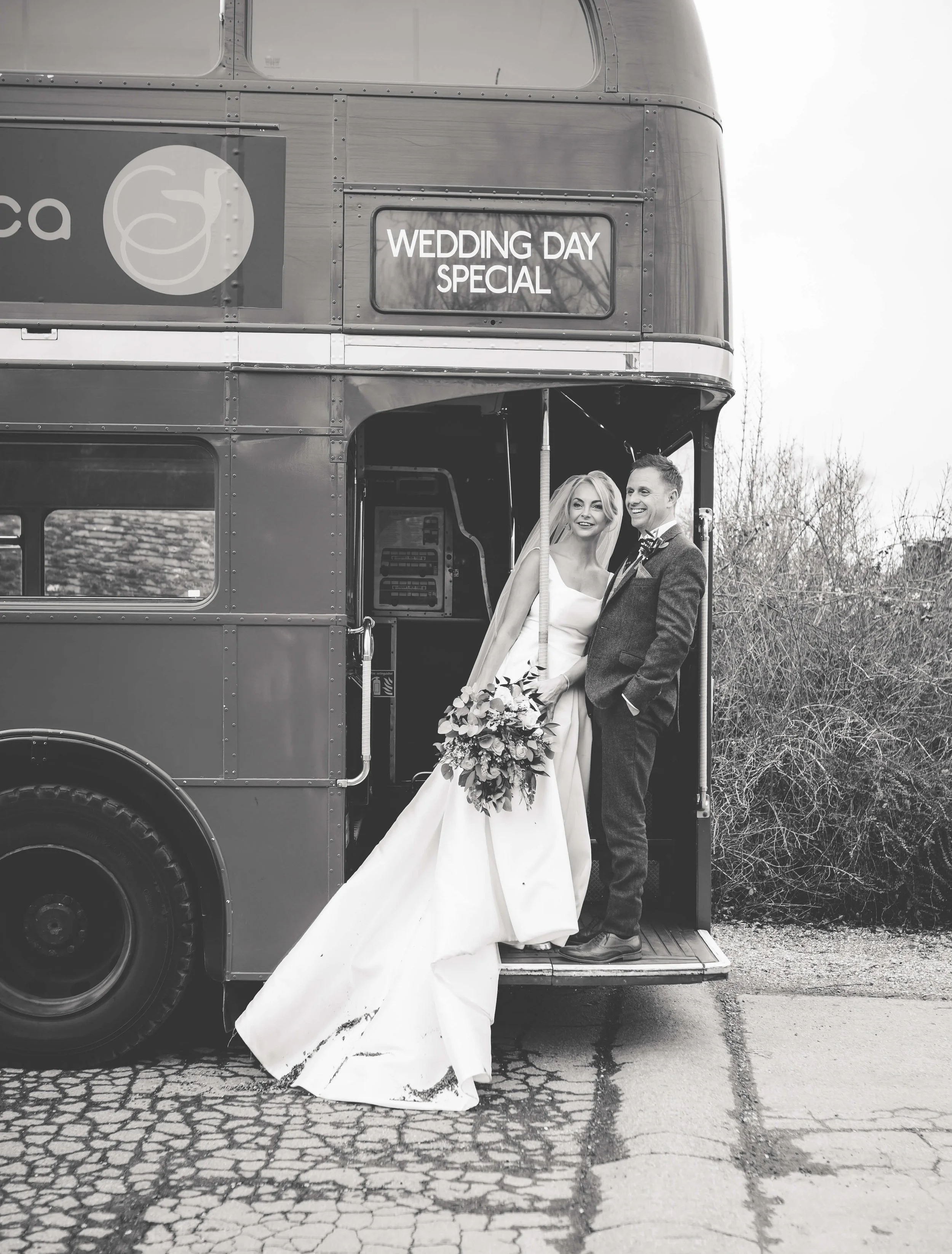 Black and white photo of a bride and groom standing at the entrance of a vintage double-decker bus, with a sign that reads 'WEDDING DAY SPECIAL', outside on a cracked pavement with leafless bushes in the background.
