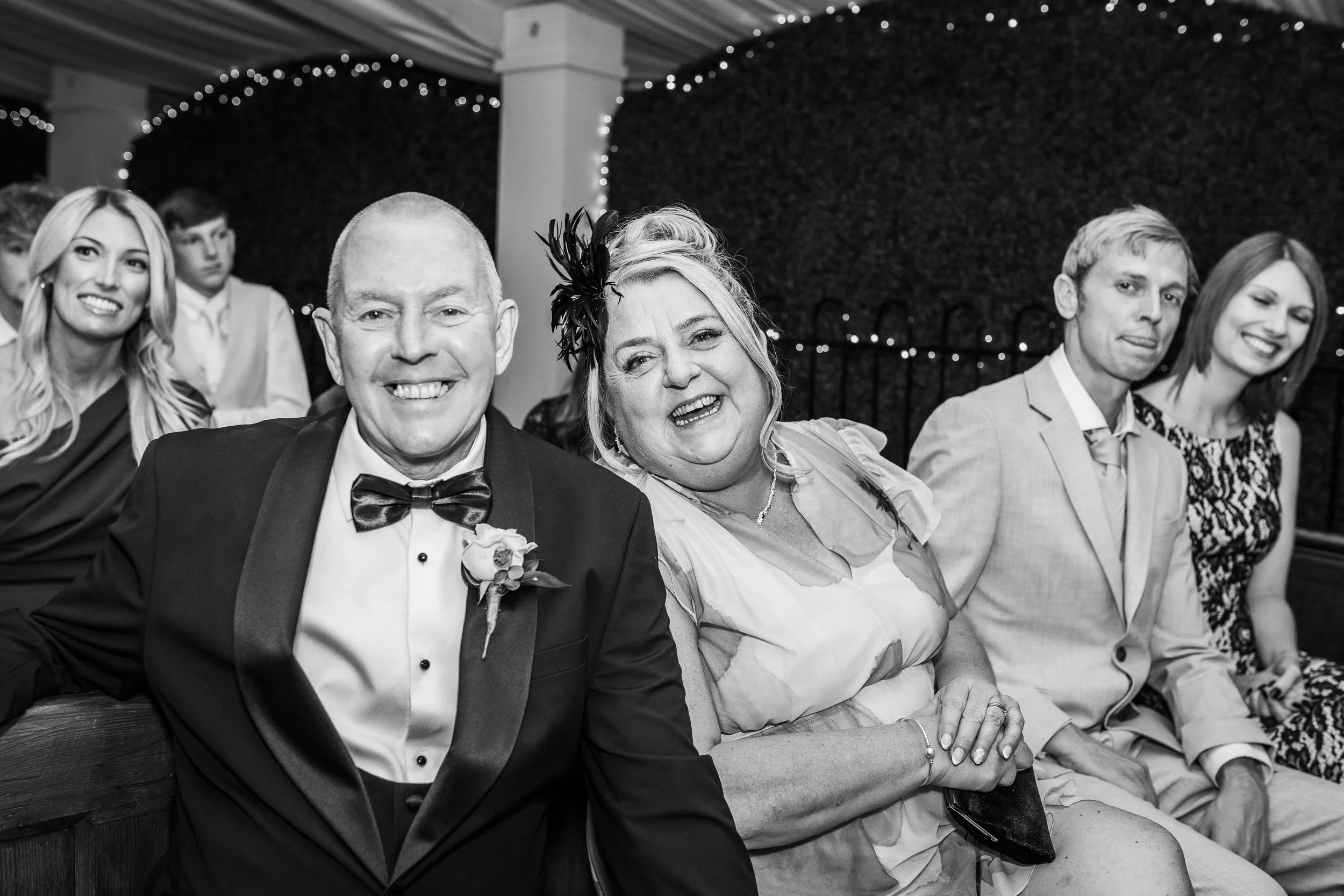 Group of people sitting at a formal event or celebration, including a man in a tuxedo and a woman with a fascinator, smiling and looking at the camera.
