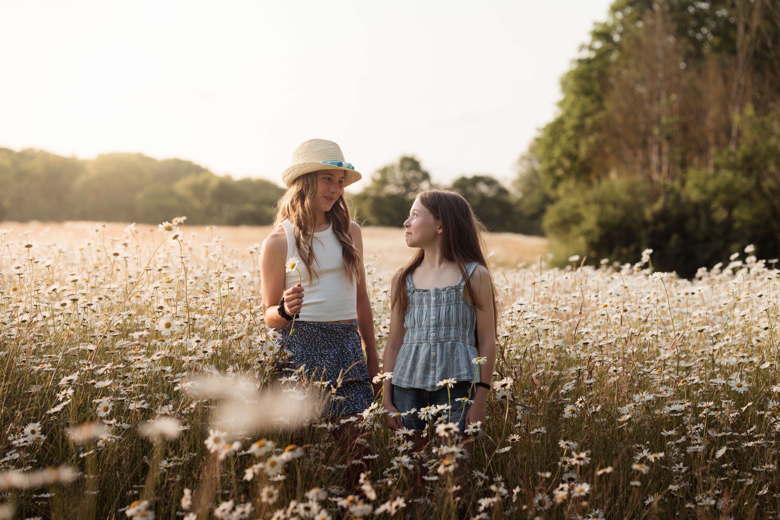 Two young girls standing in a field of daisies with trees in the background, one wearing a straw hat, holding a daisy, and looking at the other girl, who is looking back at her during sunset.